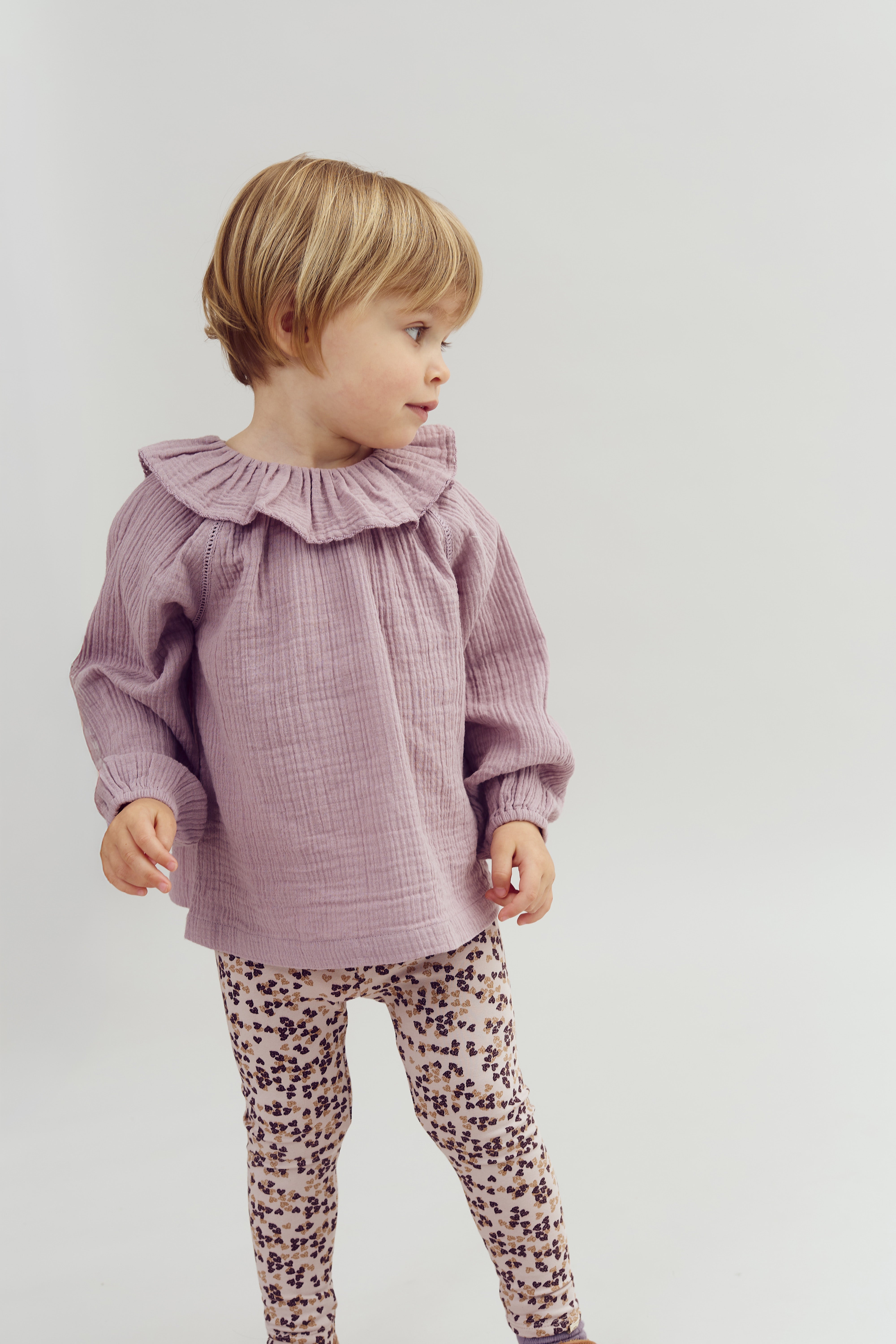 A child stands, looking to the side, wearing a purple textured top with a ruffled collar and patterned leggings, set against a simple, light background.