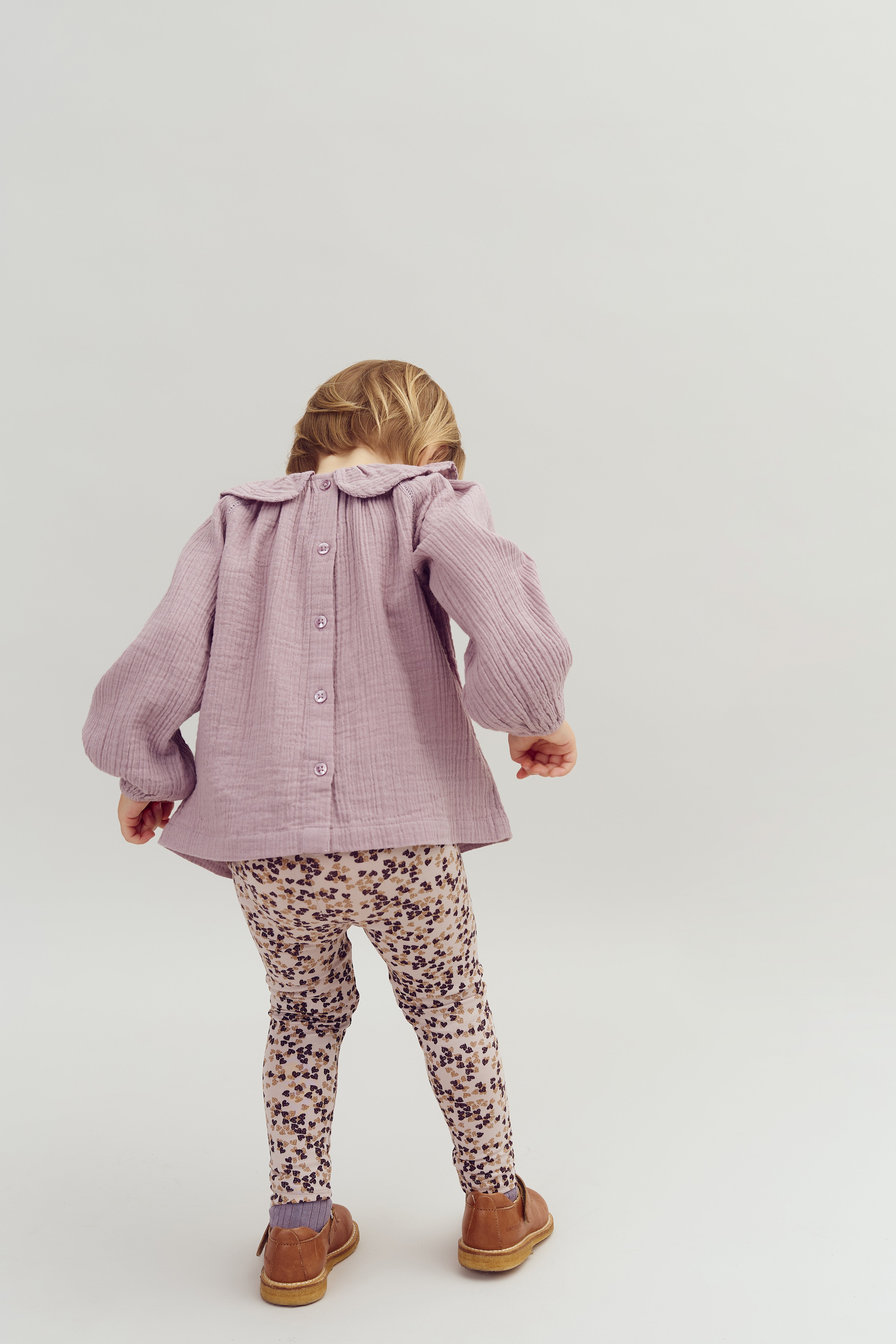 Child facing away, wearing a purple blouse and floral leggings, appears mid-step in neutral indoor setting. Blouse has buttons down the back.