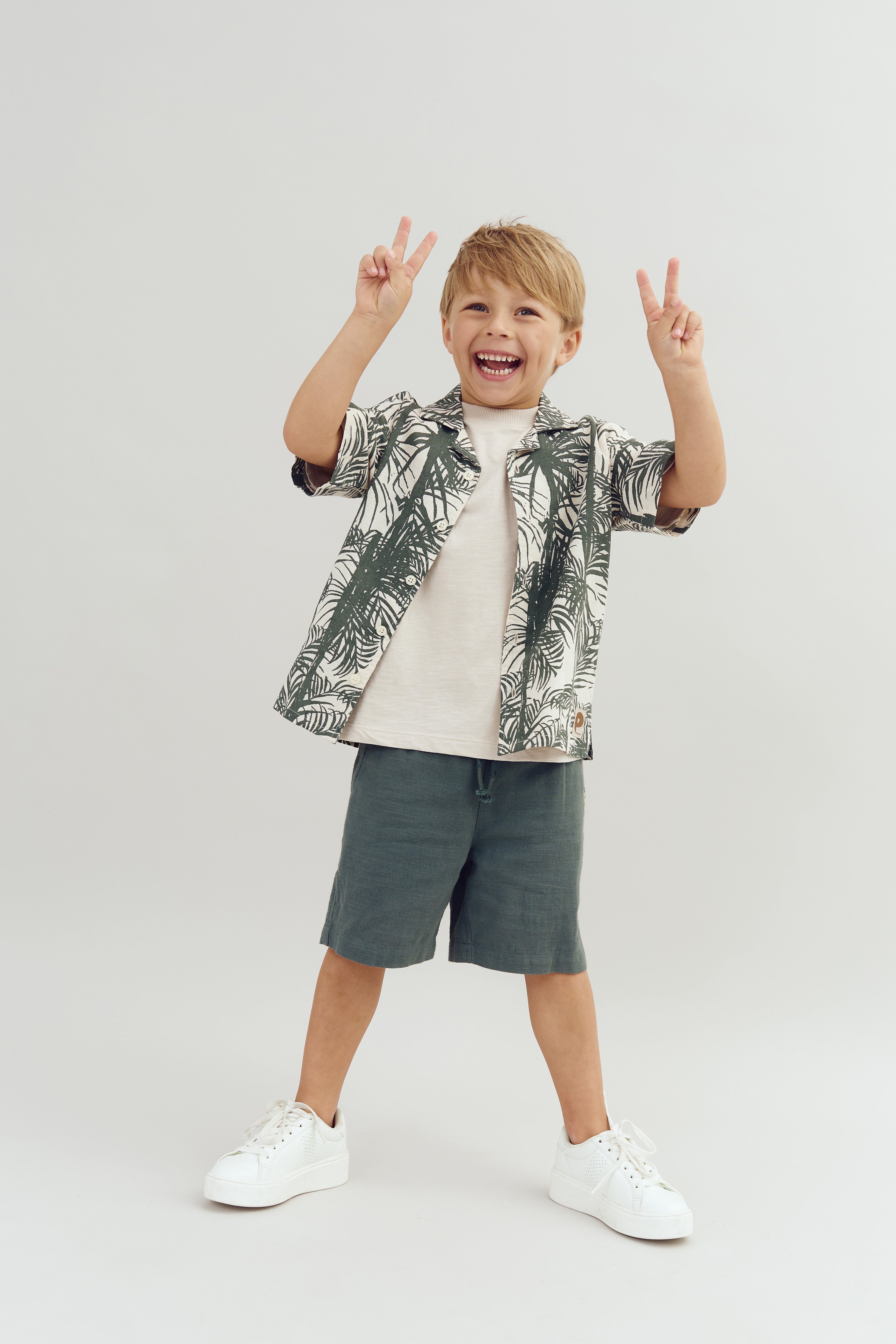 A child stands smiling, holding up two peace signs with both hands, wearing a patterned shirt and shorts against a plain background.