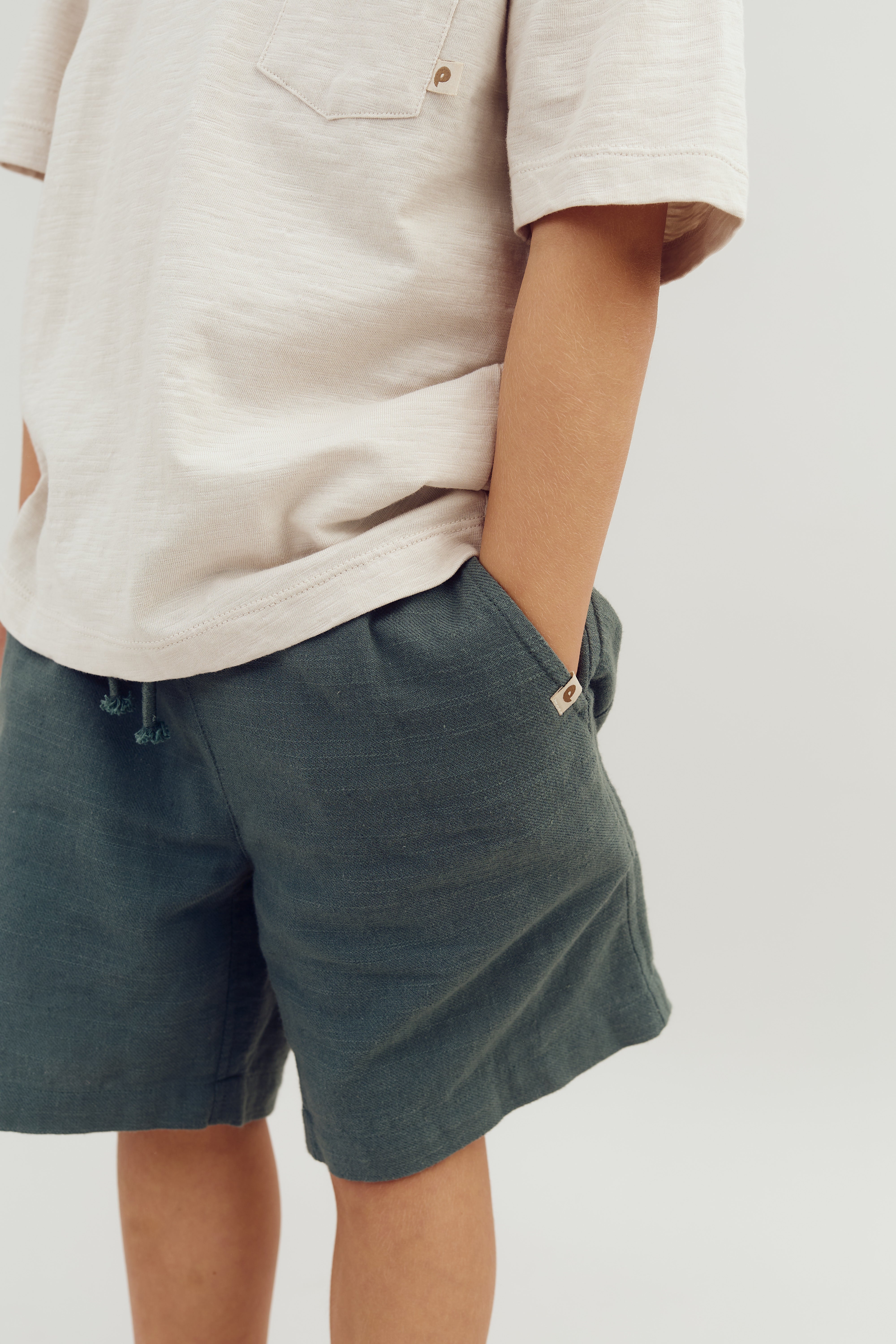 A child stands, wearing a cream short-sleeve shirt and green shorts, placing hands in pockets. The shirt has a small chest pocket and logo tag. Background is plain white.