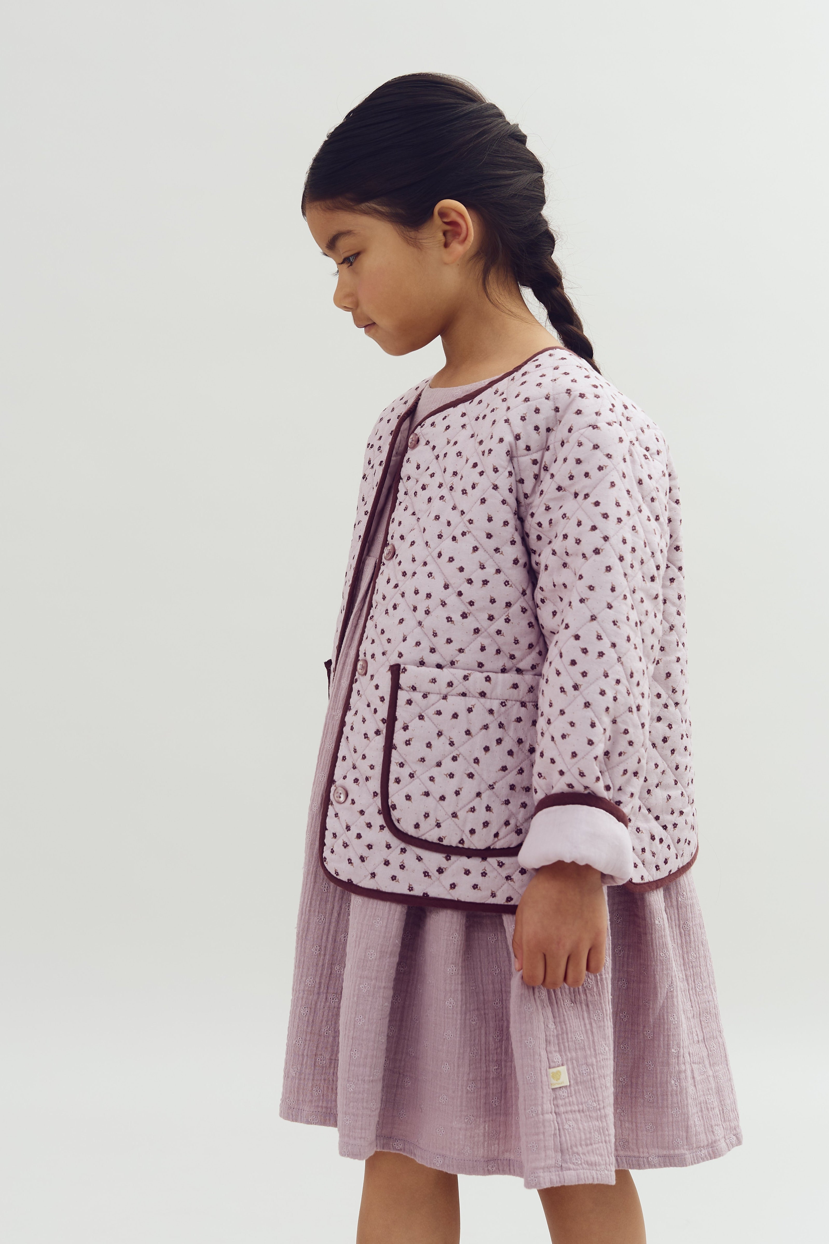 A young girl stands sideways, wearing a quilted, patterned jacket and a purple dress, with hair in a braid against a plain, light background.
