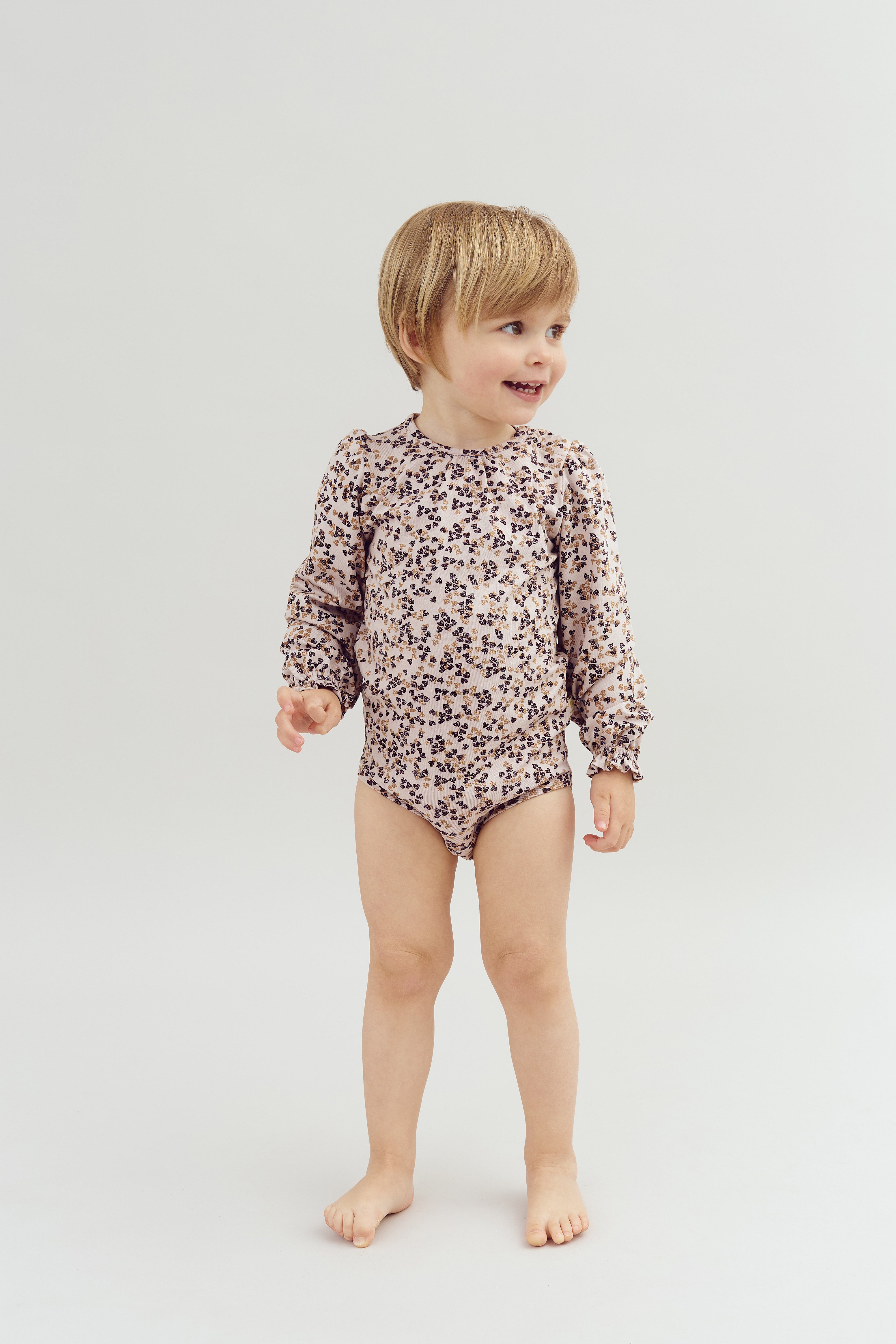 A child wearing a floral-patterned romper stands smiling on a plain white background, looking to the right with feet bare and hands relaxed.