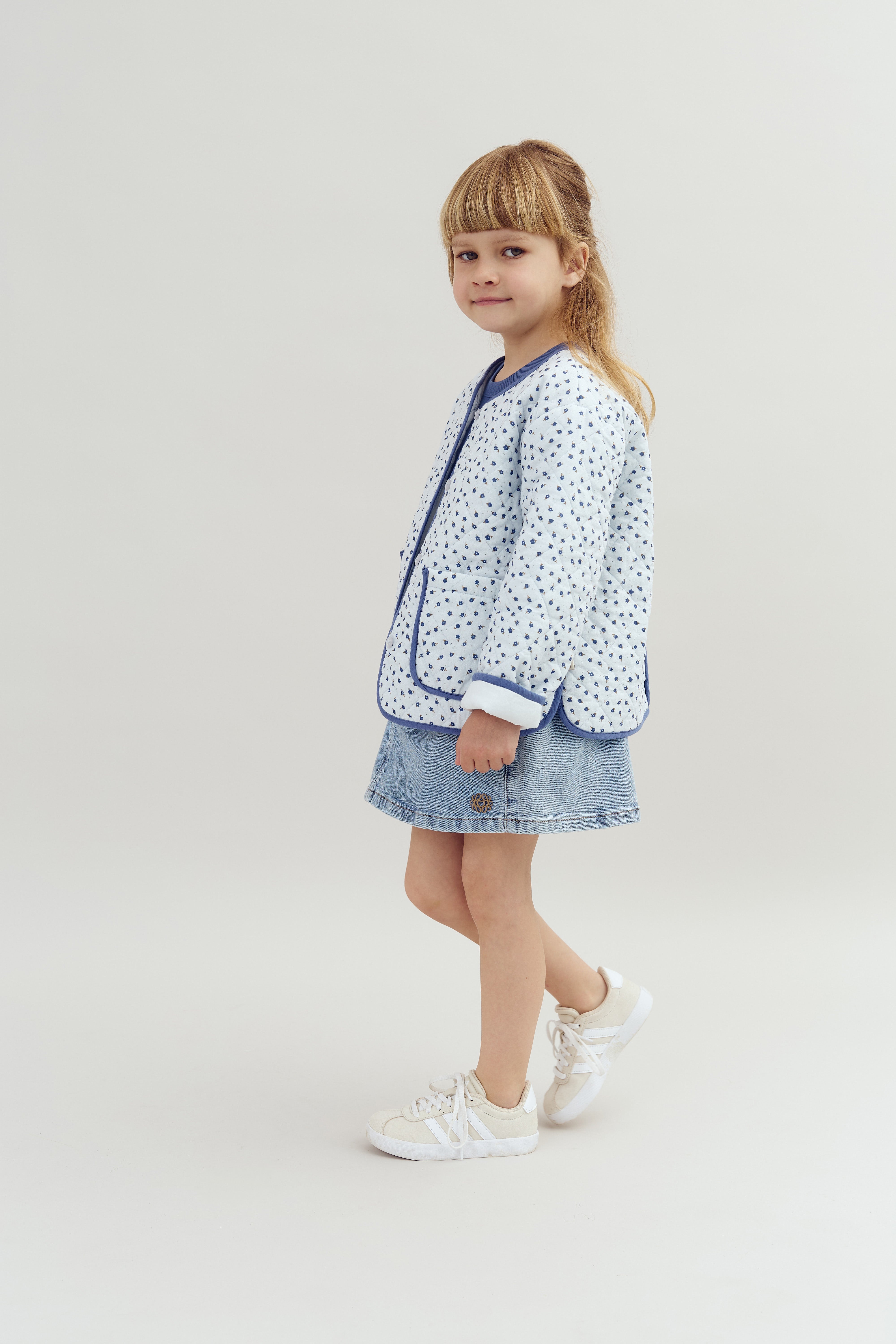 A child stands wearing a light, patterned jacket and denim skirt, paired with white sneakers. The background is a plain, light-colored studio setting.