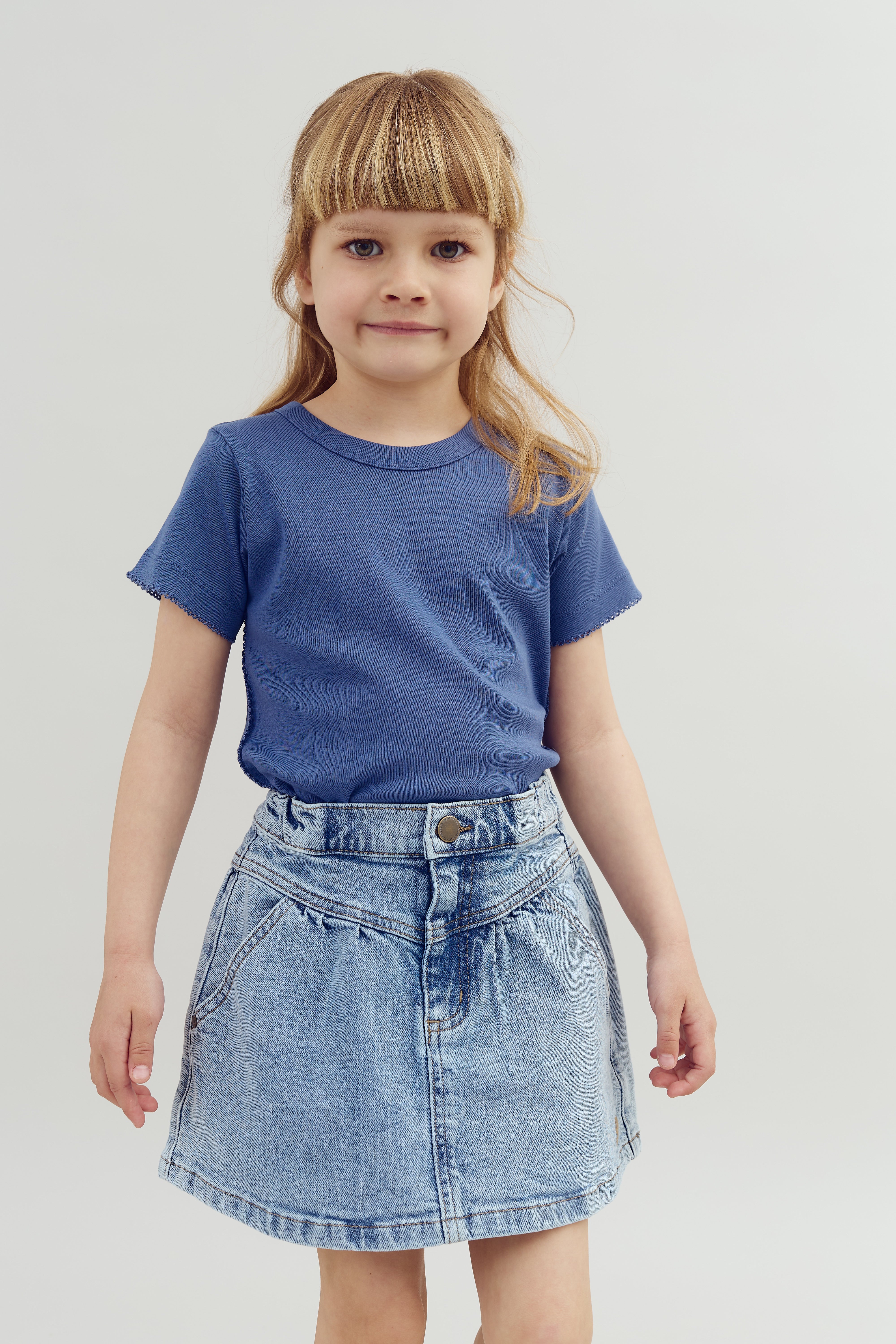 A young girl stands, wearing a blue T-shirt and denim skirt, against a plain white background. She has straight hair with bangs and is looking forward.