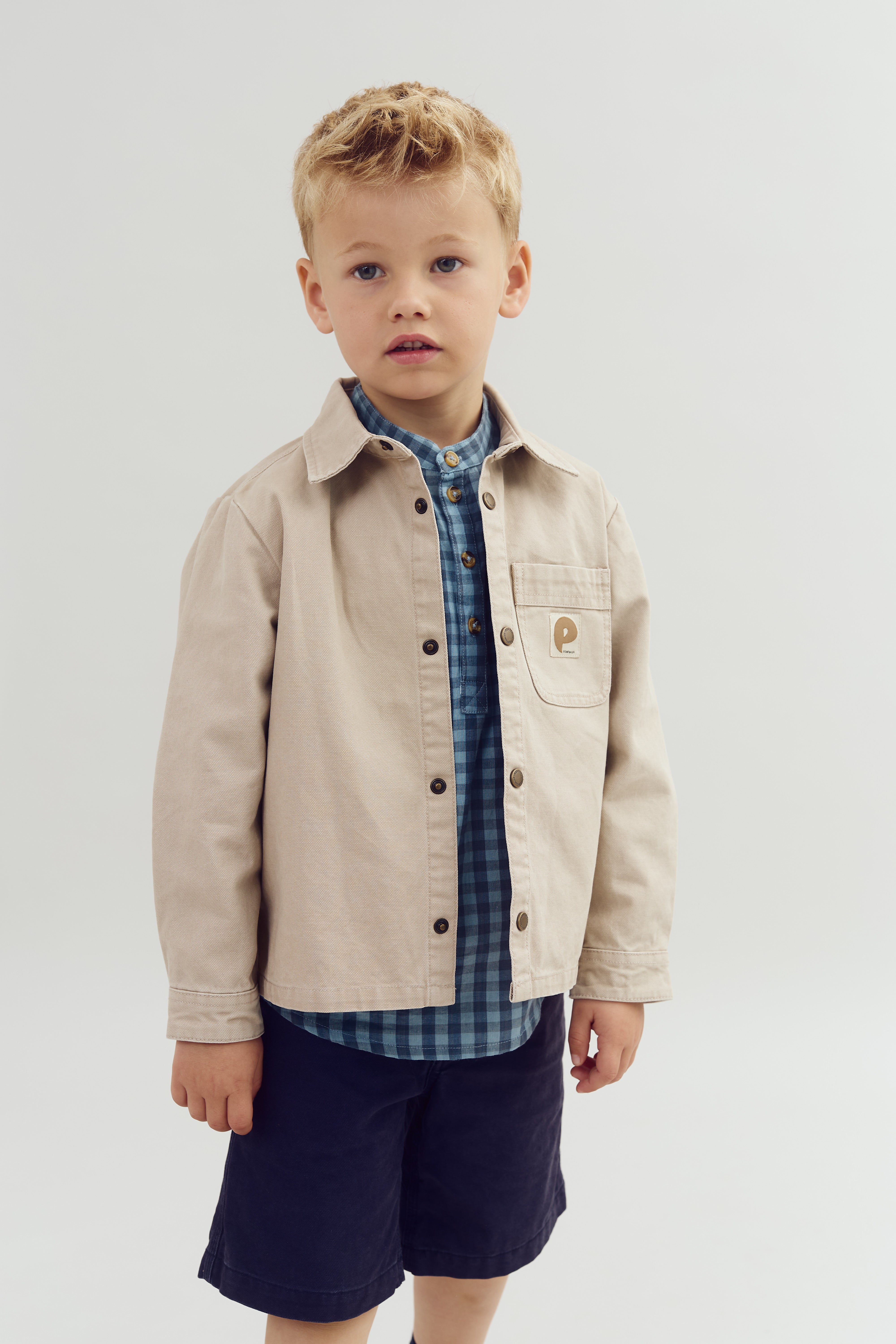 A young boy stands in a photo studio. He wears a beige jacket with a pocket labeled "P" and a blue checkered shirt, paired with navy shorts.