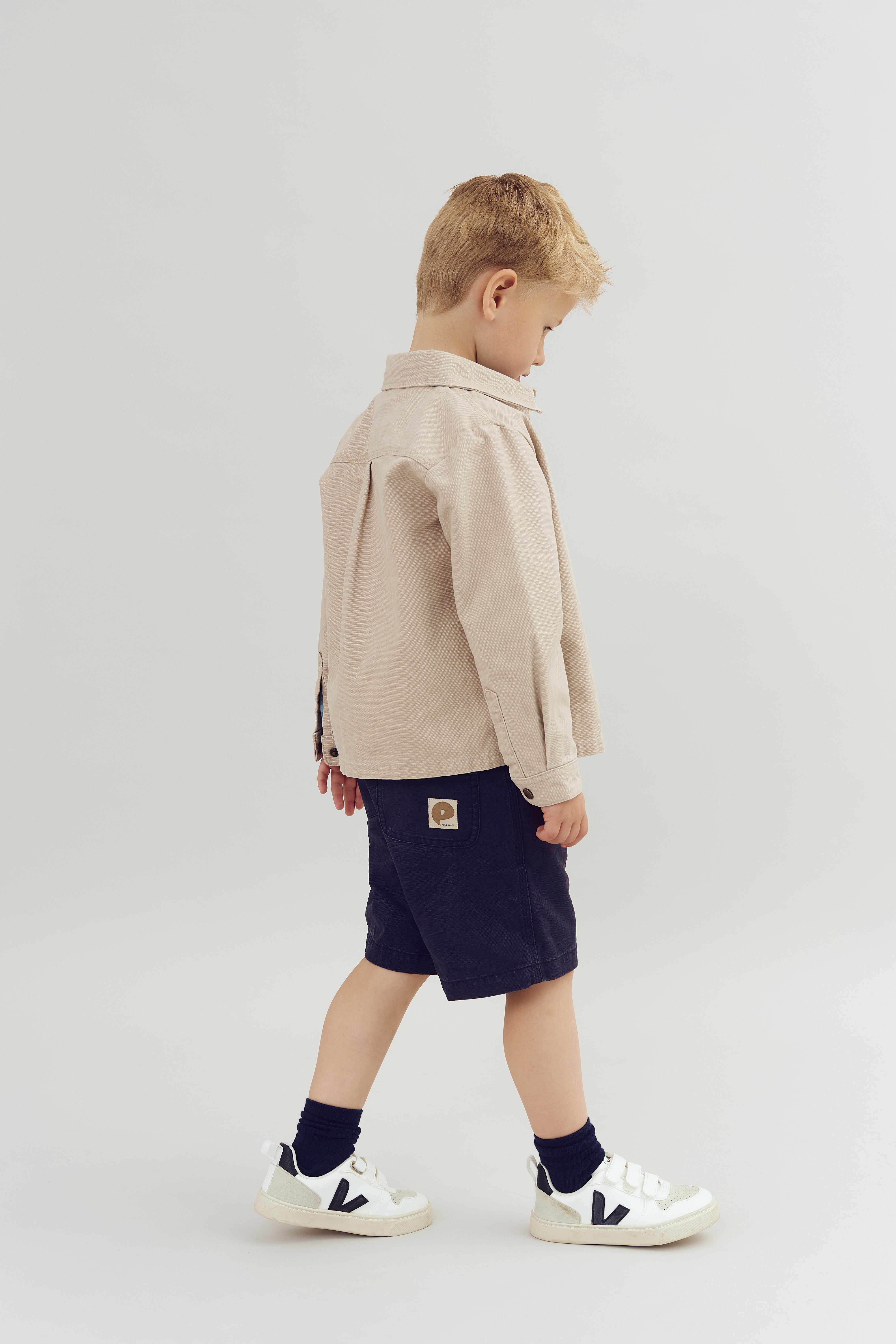 A young child in a beige jacket and dark shorts stands sideways, focusing downward. The background is a plain white studio setting. Clothing features a visible "P" logo on shorts.