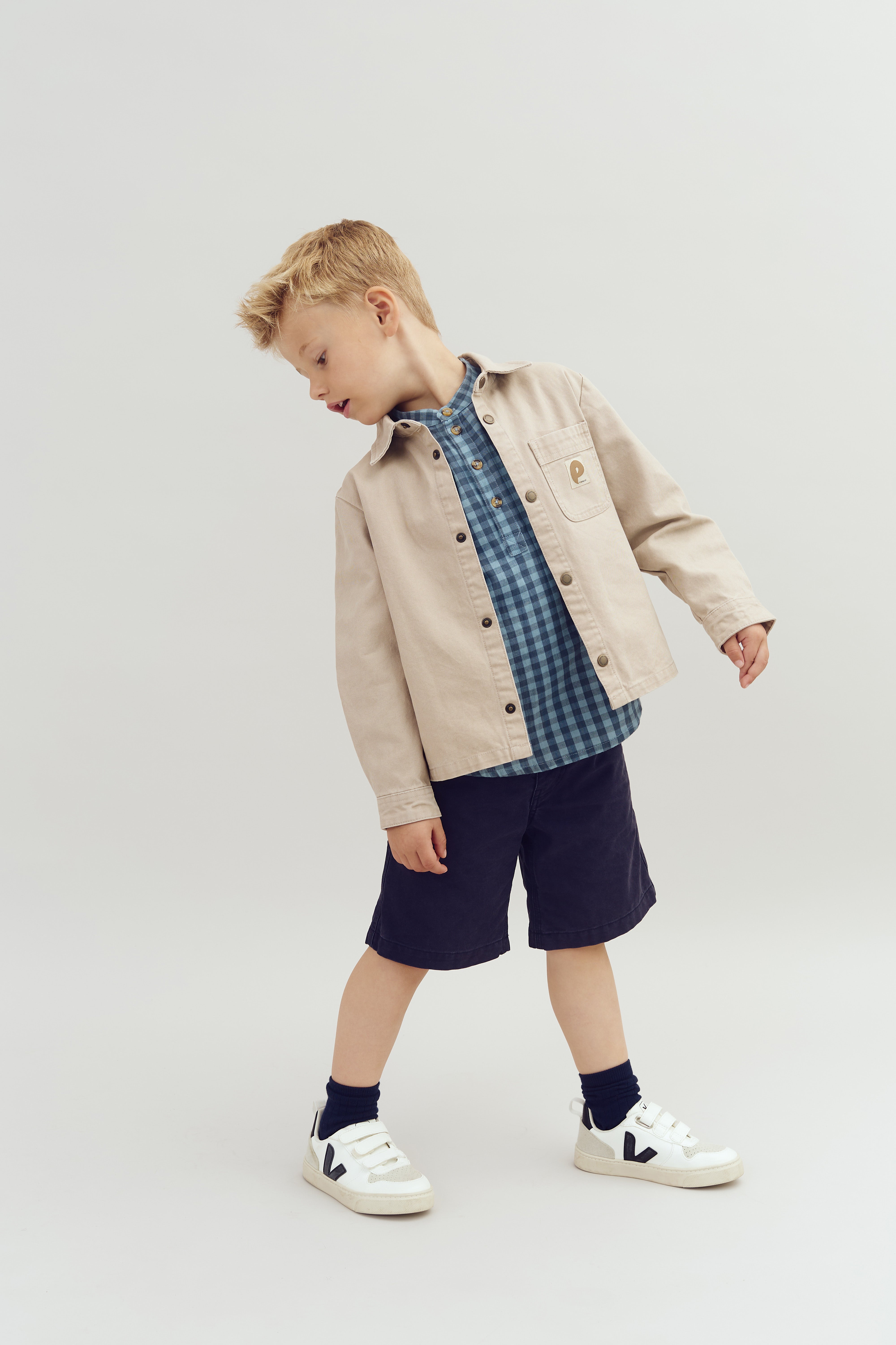 A child, wearing a beige jacket, blue checkered shirt, and dark shorts, looks down to the side while standing on a plain background.