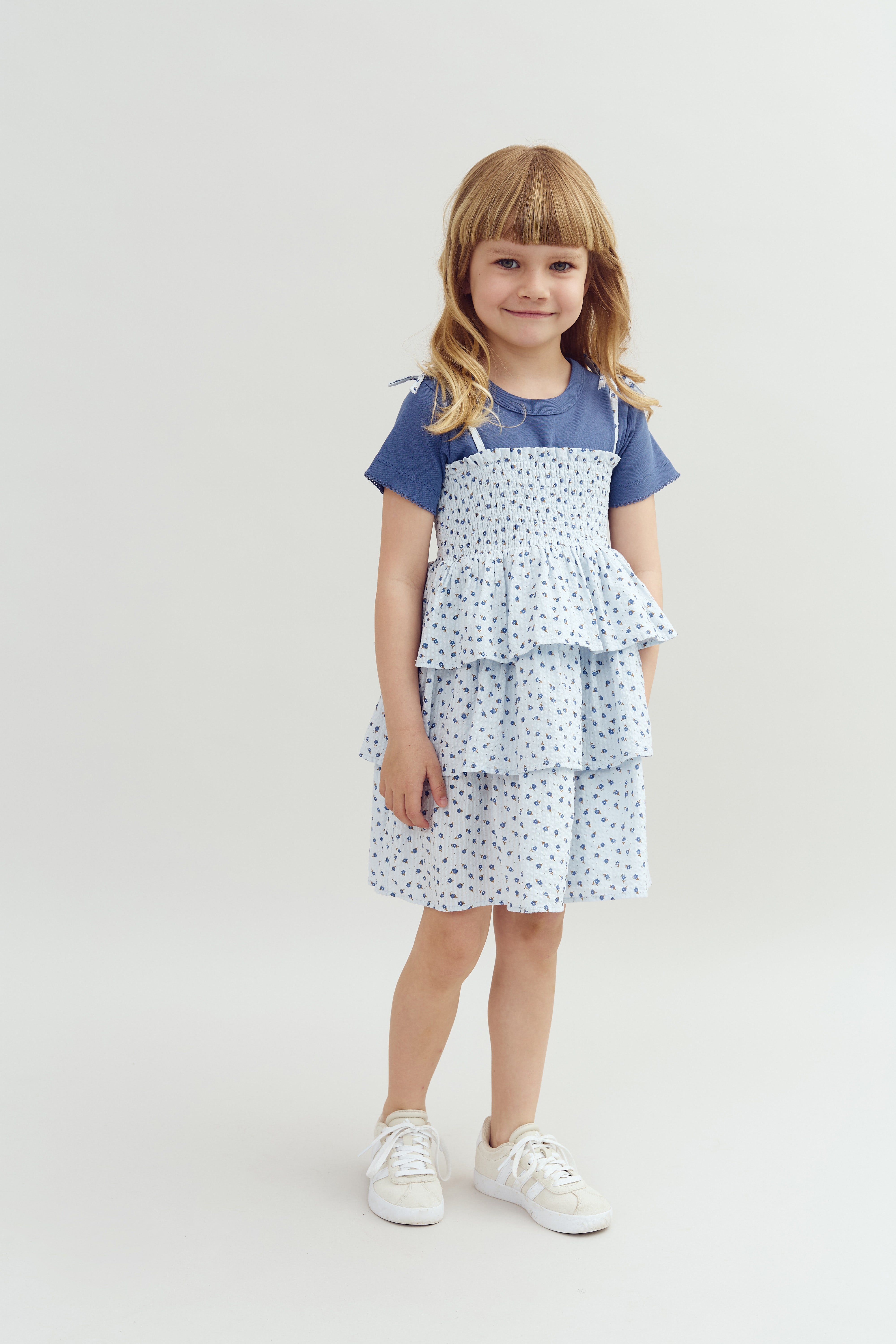 A young girl stands smiling, wearing a blue patterned dress with layered ruffles and white sneakers, against a plain white background.