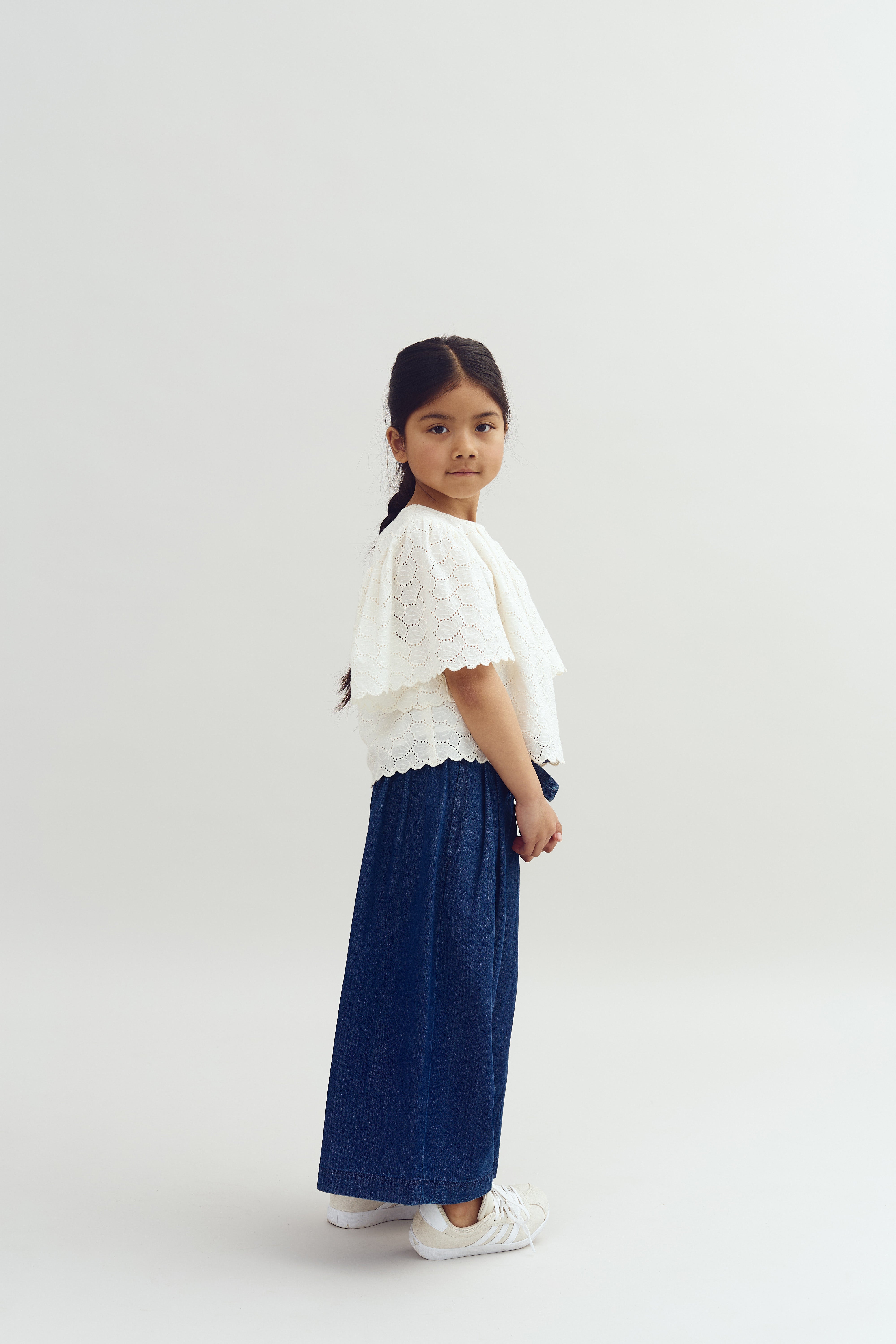 A young girl stands in profile, wearing a white lace top and blue skirt, against a plain, light background.