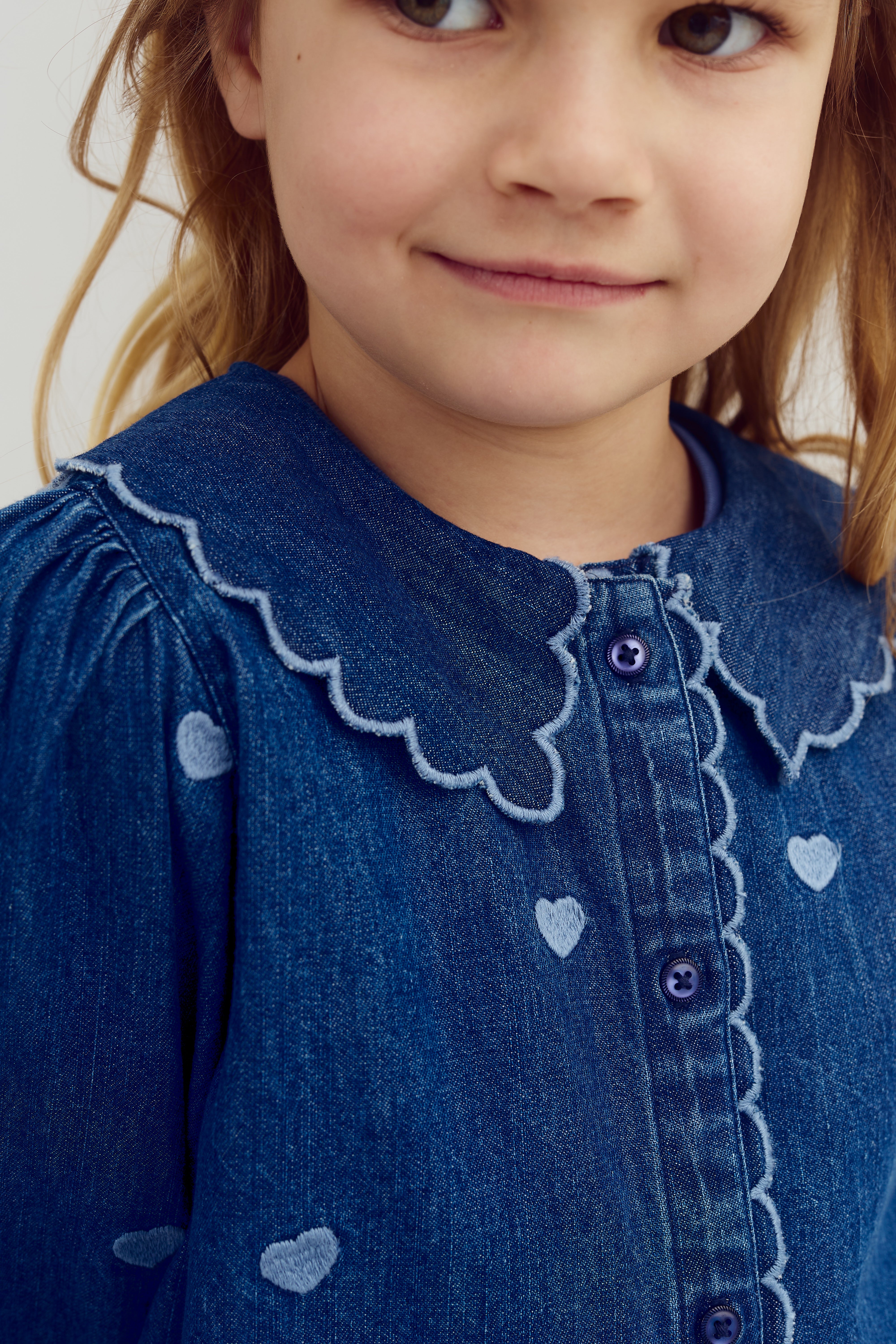 A child in a blue denim dress with heart patterns stands against a plain background. The dress features scalloped edges and buttons down the front.