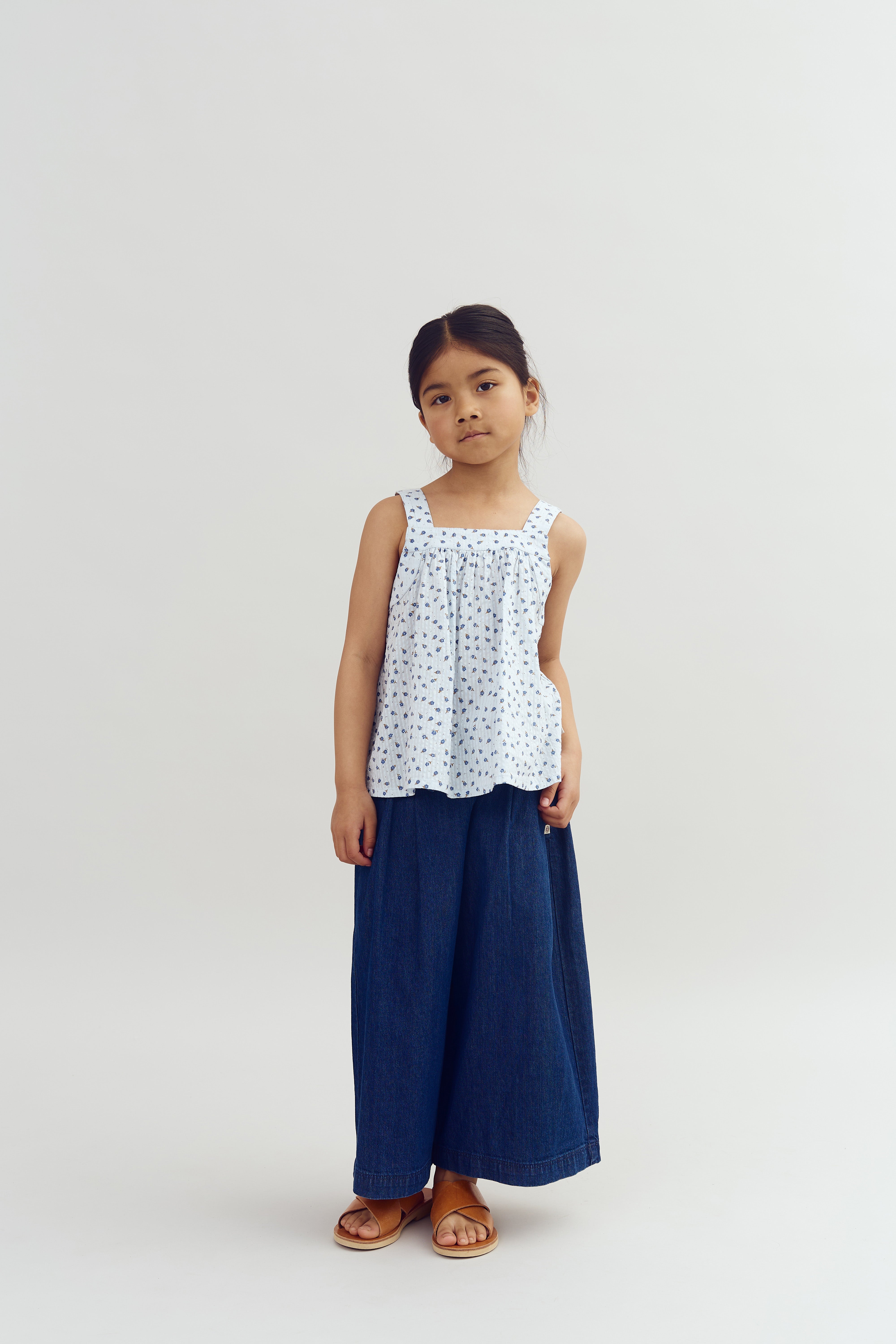 A young girl stands, wearing a white, patterned sleeveless top and wide-leg denim pants, posing calmly against a plain, neutral background.