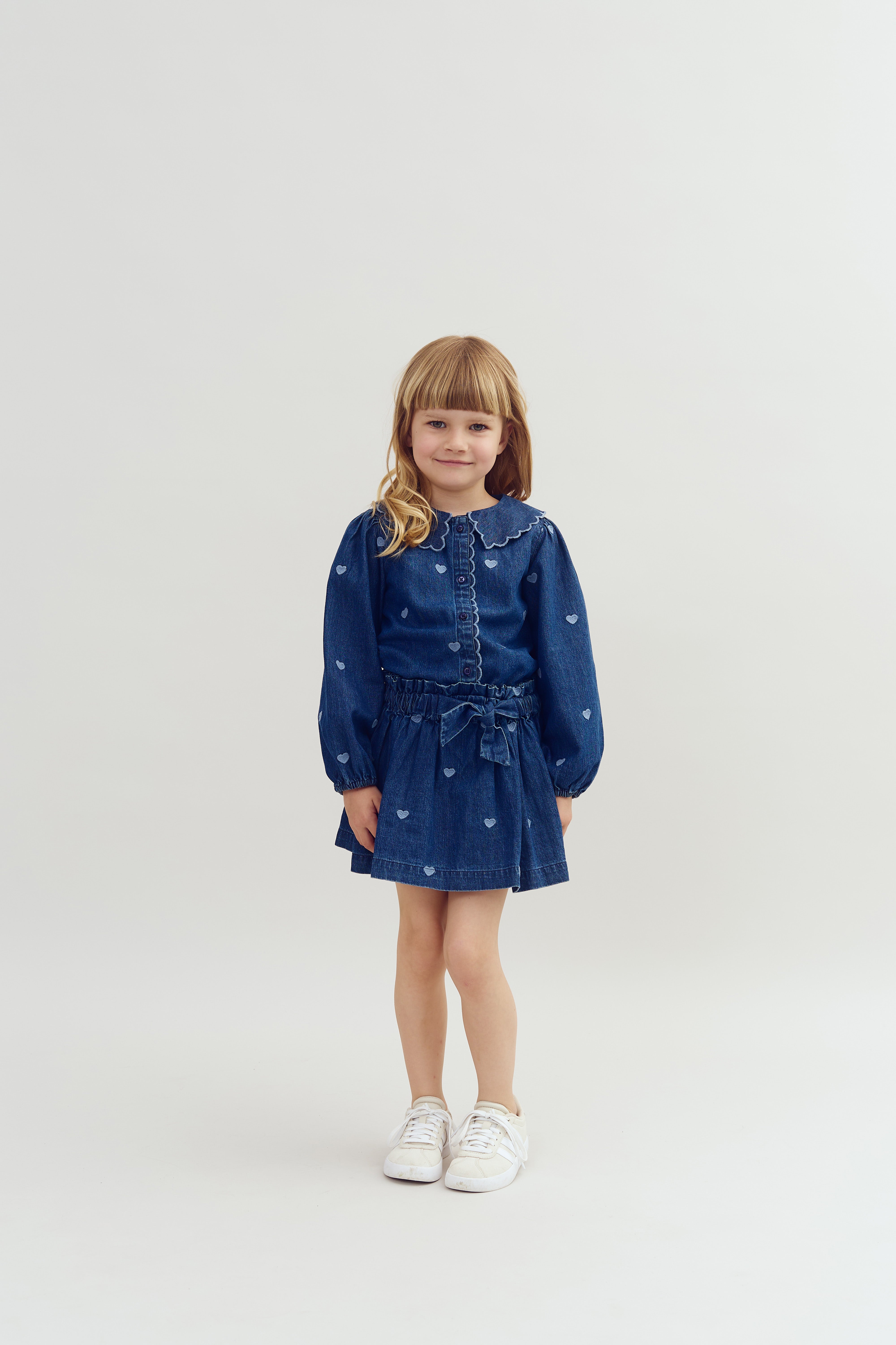 A child stands wearing a blue denim dress with heart patterns, hands by her side, in a plain, neutral background. She is also wearing white sneakers.