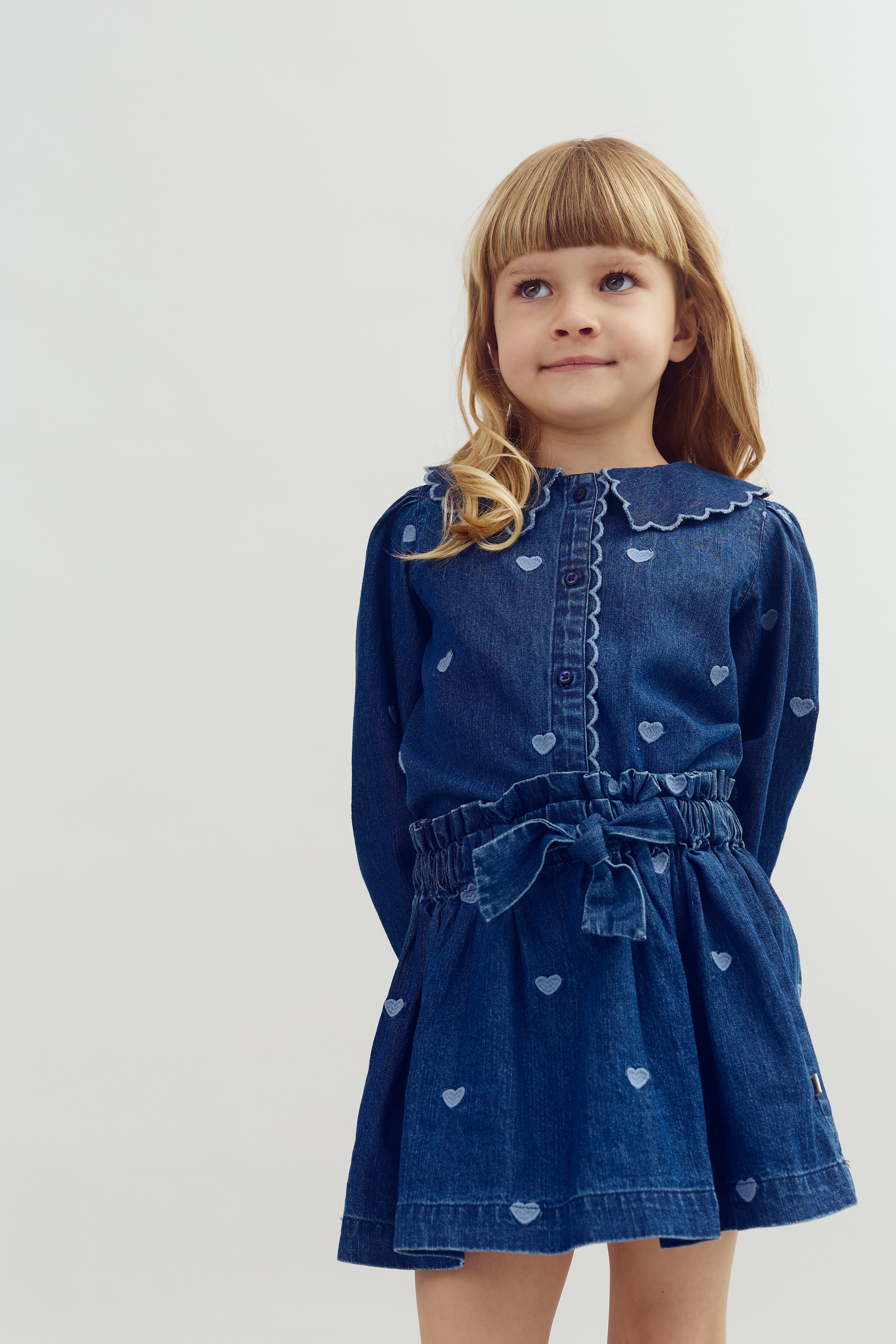 A young child wearing a blue, heart-patterned denim dress stands against a plain, light background, looking upwards with a gentle smile.
