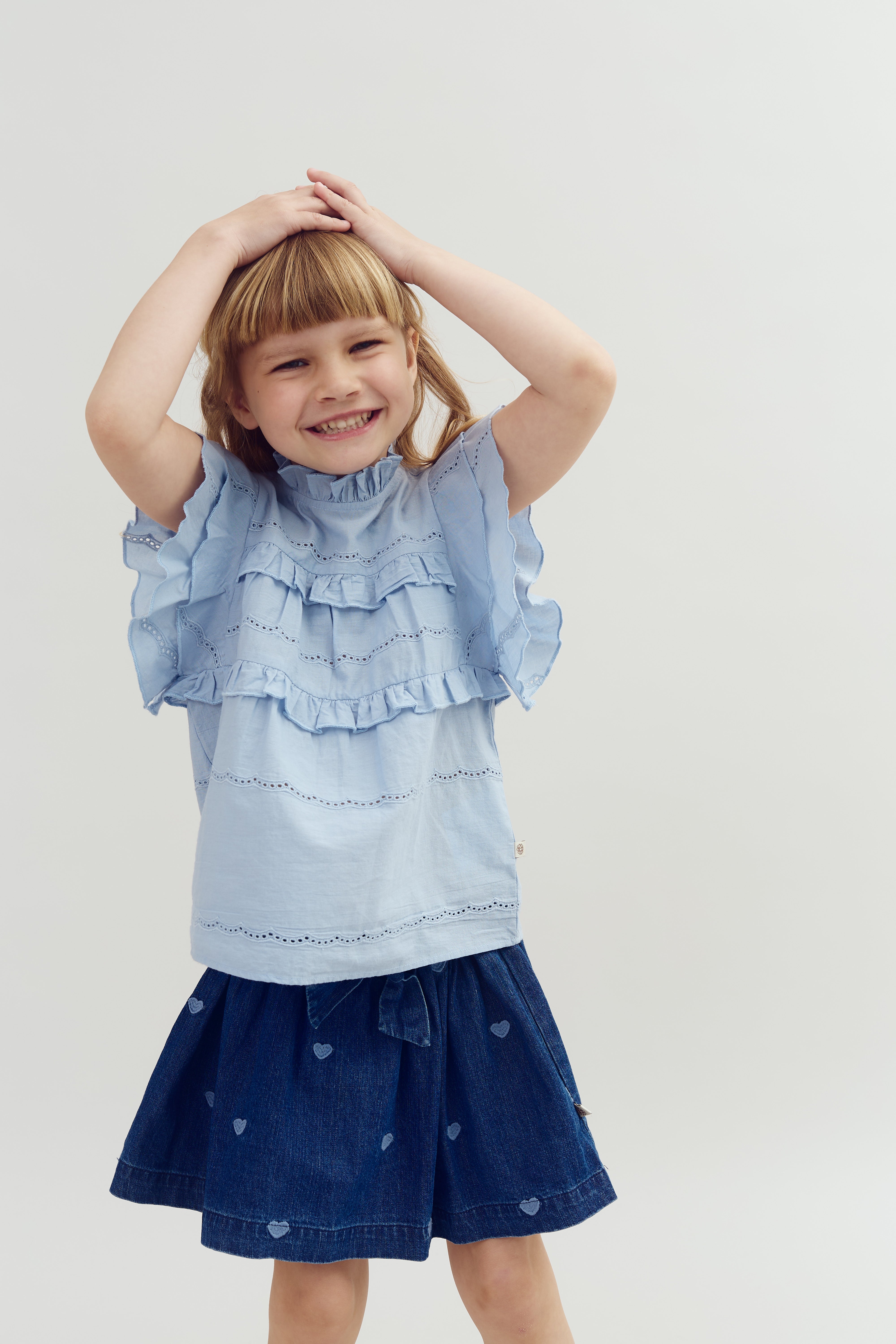 Child smiling, hands on head, wearing a ruffled blue blouse and a denim skirt with heart patterns, against a plain white background.