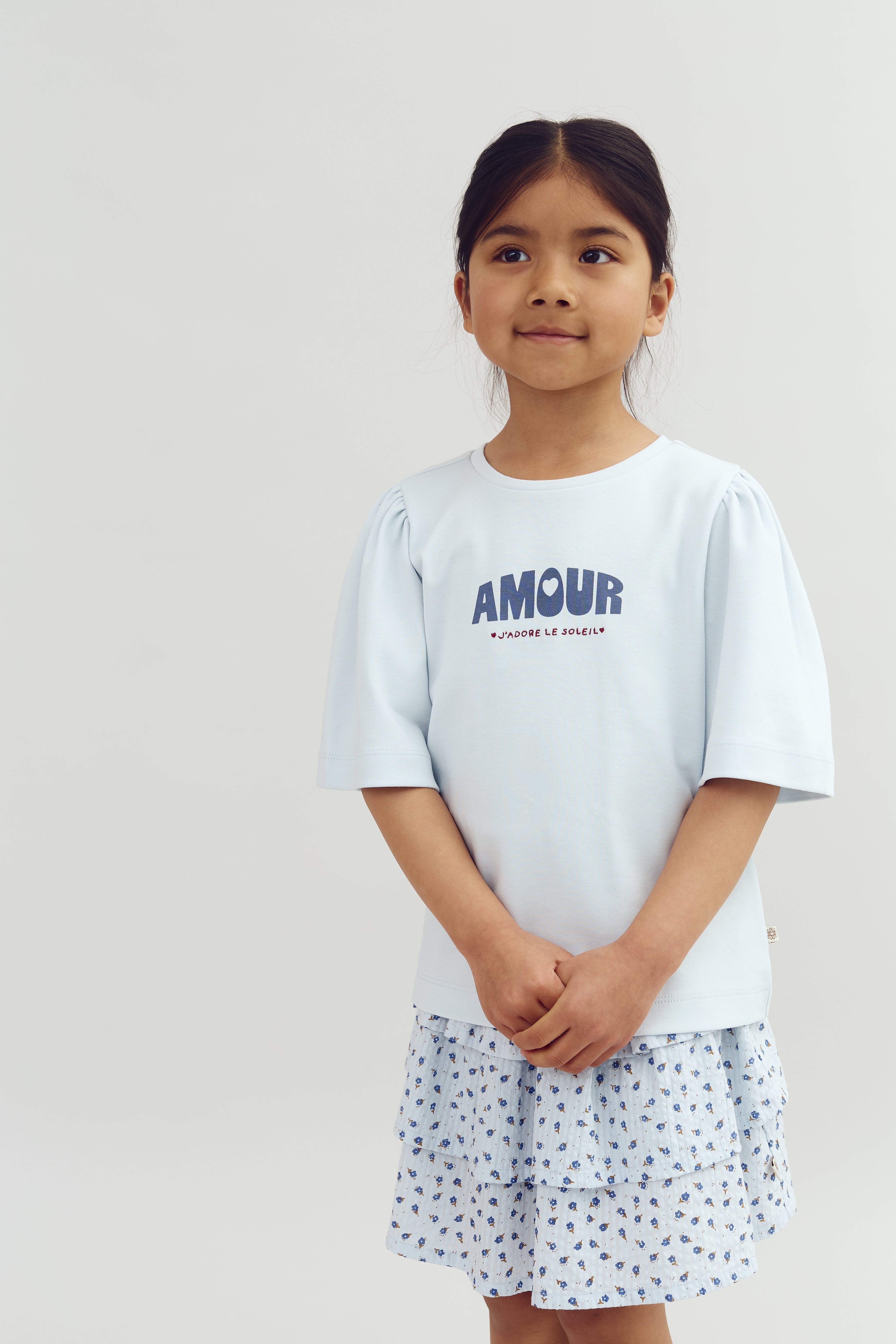 A girl stands against a plain background, wearing a light blue shirt with "AMOUR J'adore le soleil" printed on it and a tiered floral-patterned skirt, hands clasped.