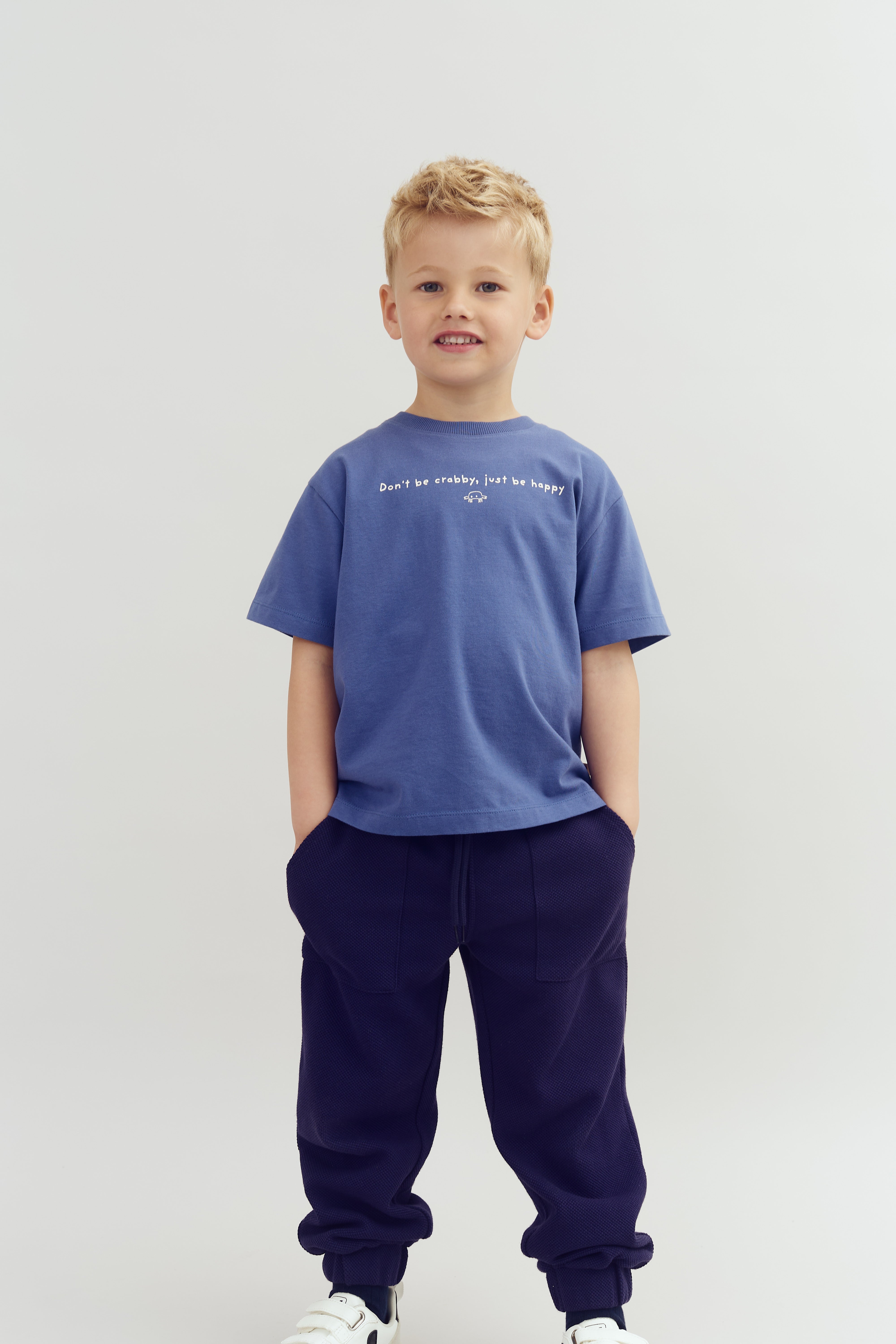 Child wearing a blue shirt with "Don't be crabby, just be happy" text, standing with hands in pockets, dressed in dark pants and white sneakers, against a plain background.