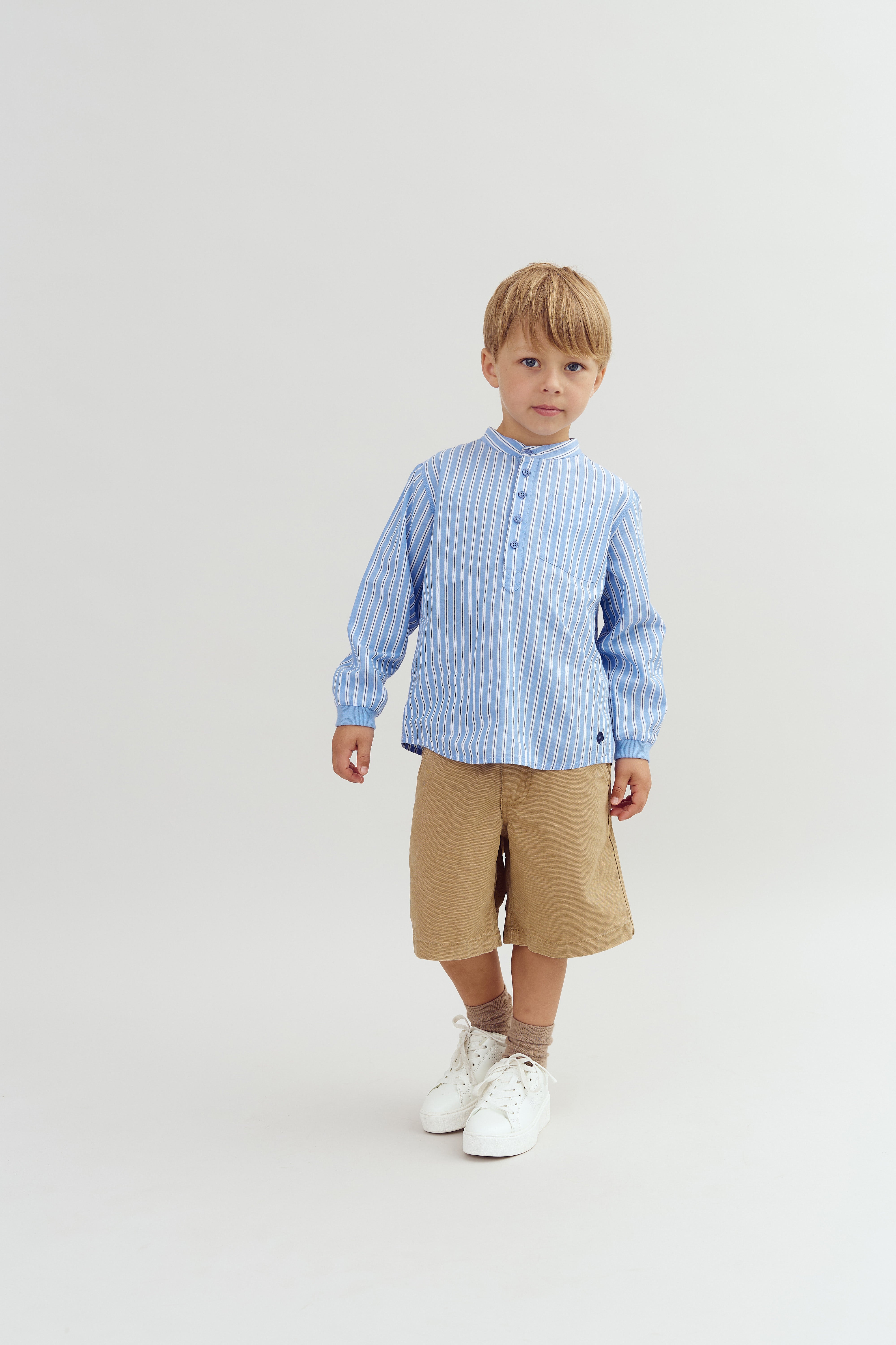 A child stands calmly wearing a blue striped shirt, beige shorts, and white sneakers against a plain white background. The setting is minimal and clean.