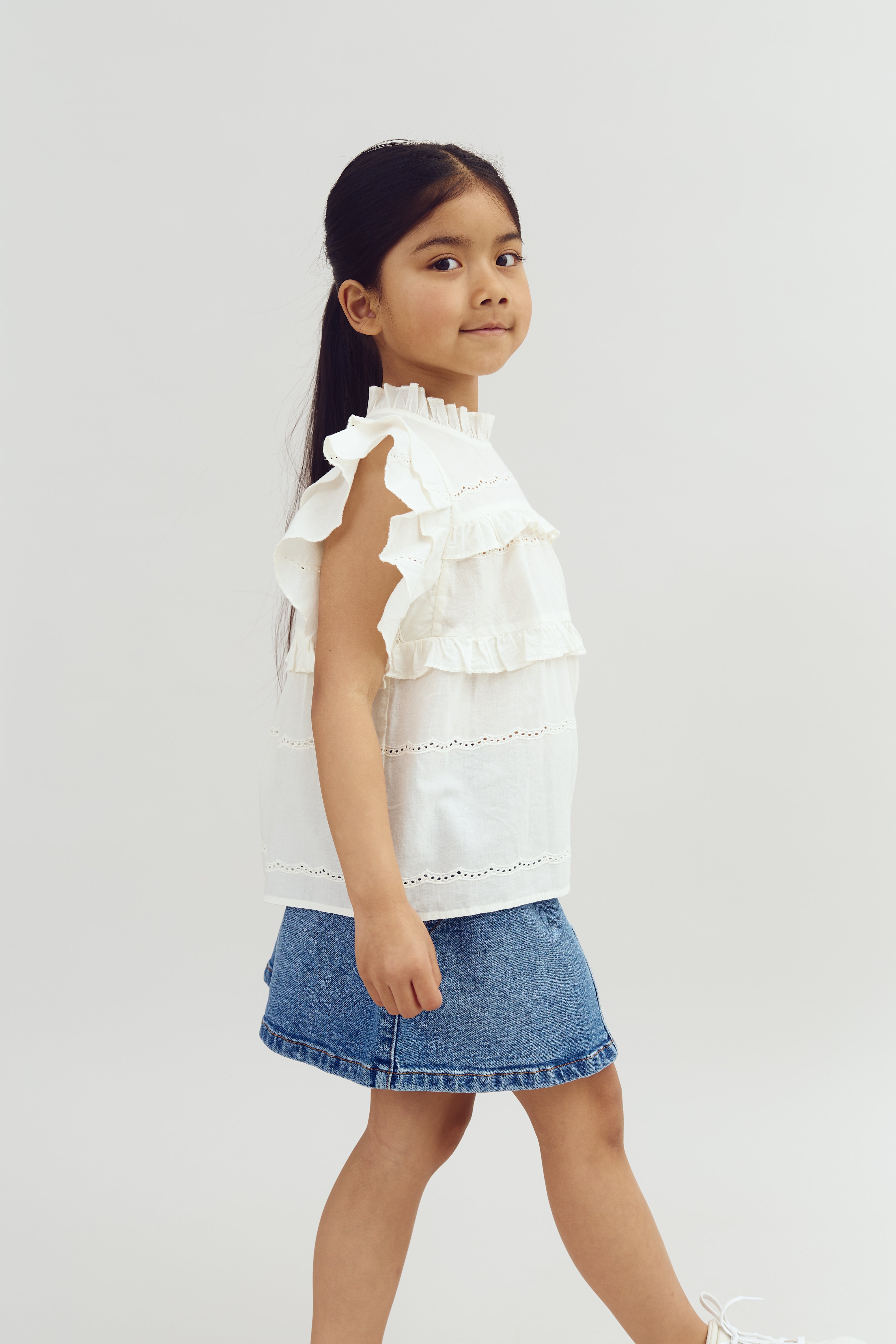 A young child in a white ruffled blouse and denim skirt walks confidently against a plain, light background, conveying a sense of motion and style.
