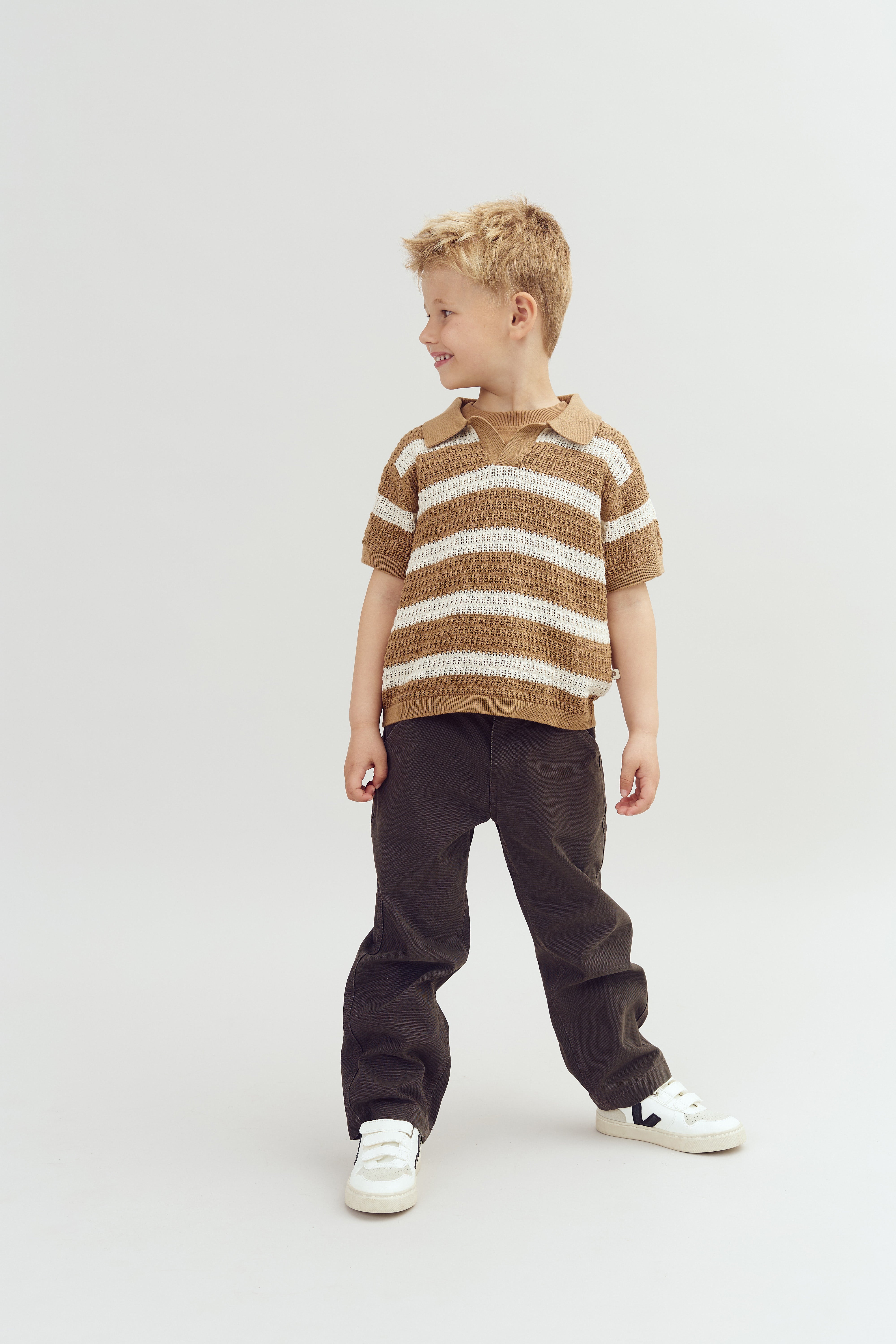 A child, standing and smiling to the side, wears a brown-and-white striped sweater, dark pants, and white sneakers. The background is plain and light-colored.