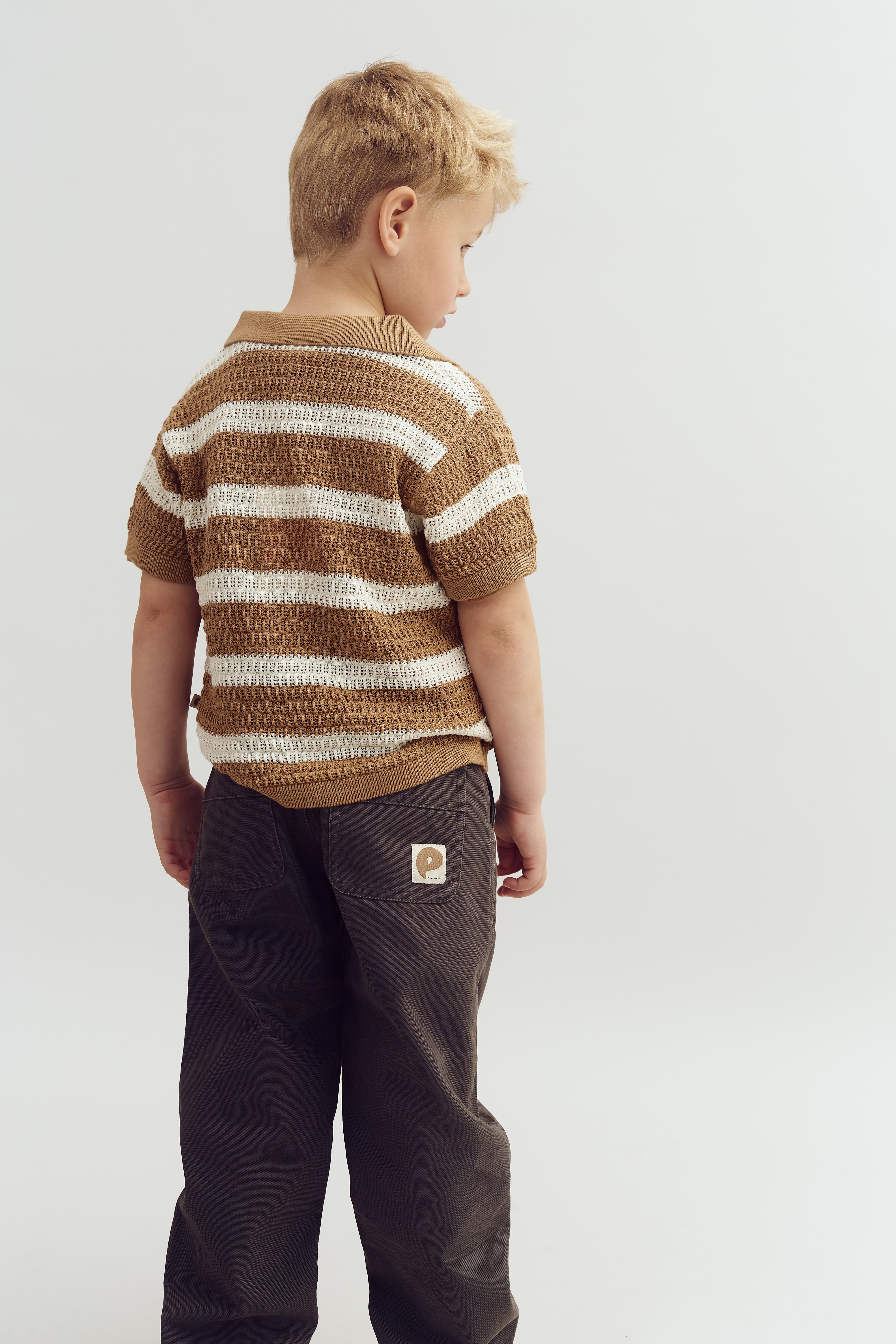 A child stands facing away, wearing a brown and white striped shirt and dark pants with a logo on the back pocket, in a plain studio setting.