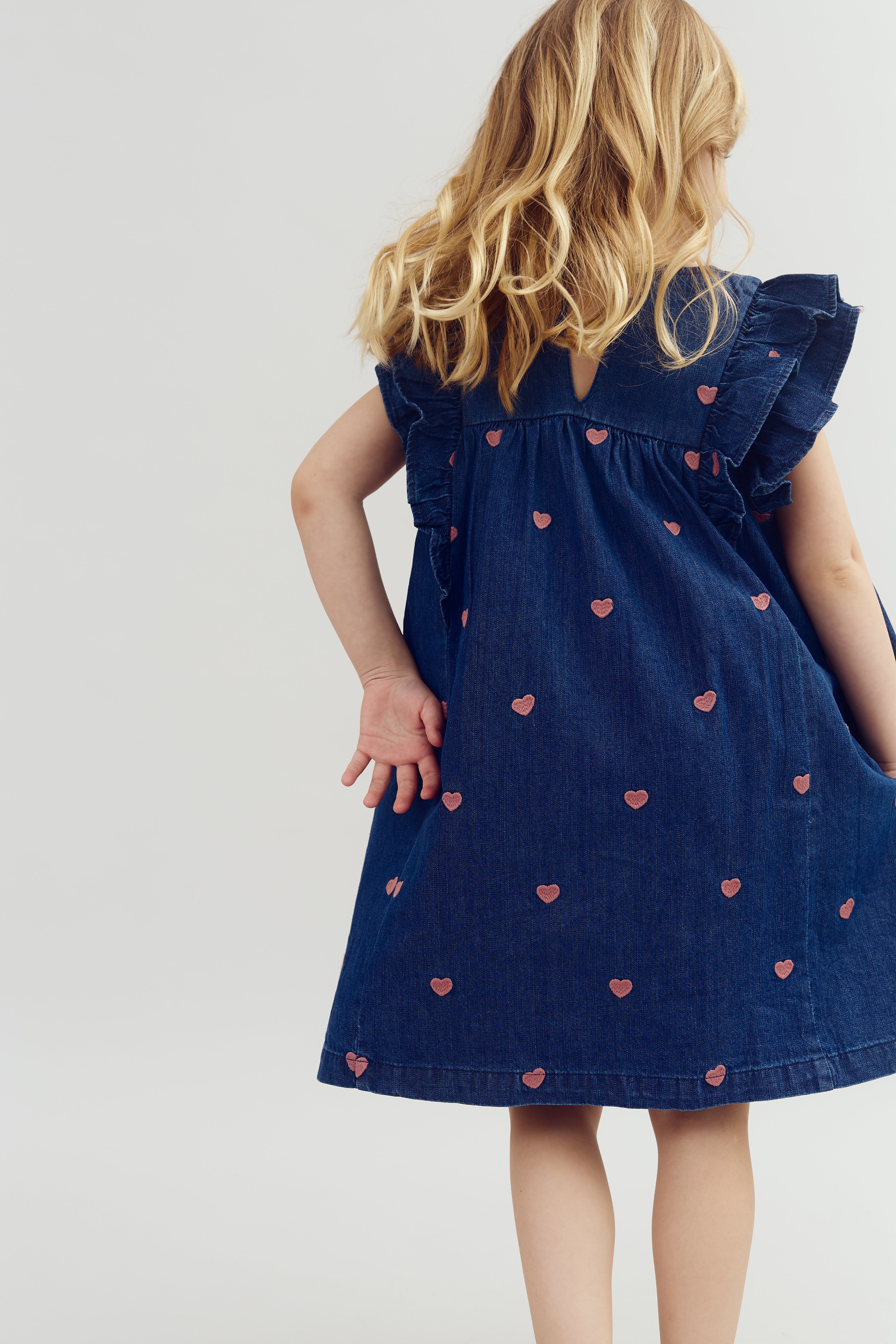 Child wearing a blue dress adorned with small pink hearts, standing with one hand behind back in a neutral-toned studio setting. The dress has ruffled sleeves, and hair is light brown.
