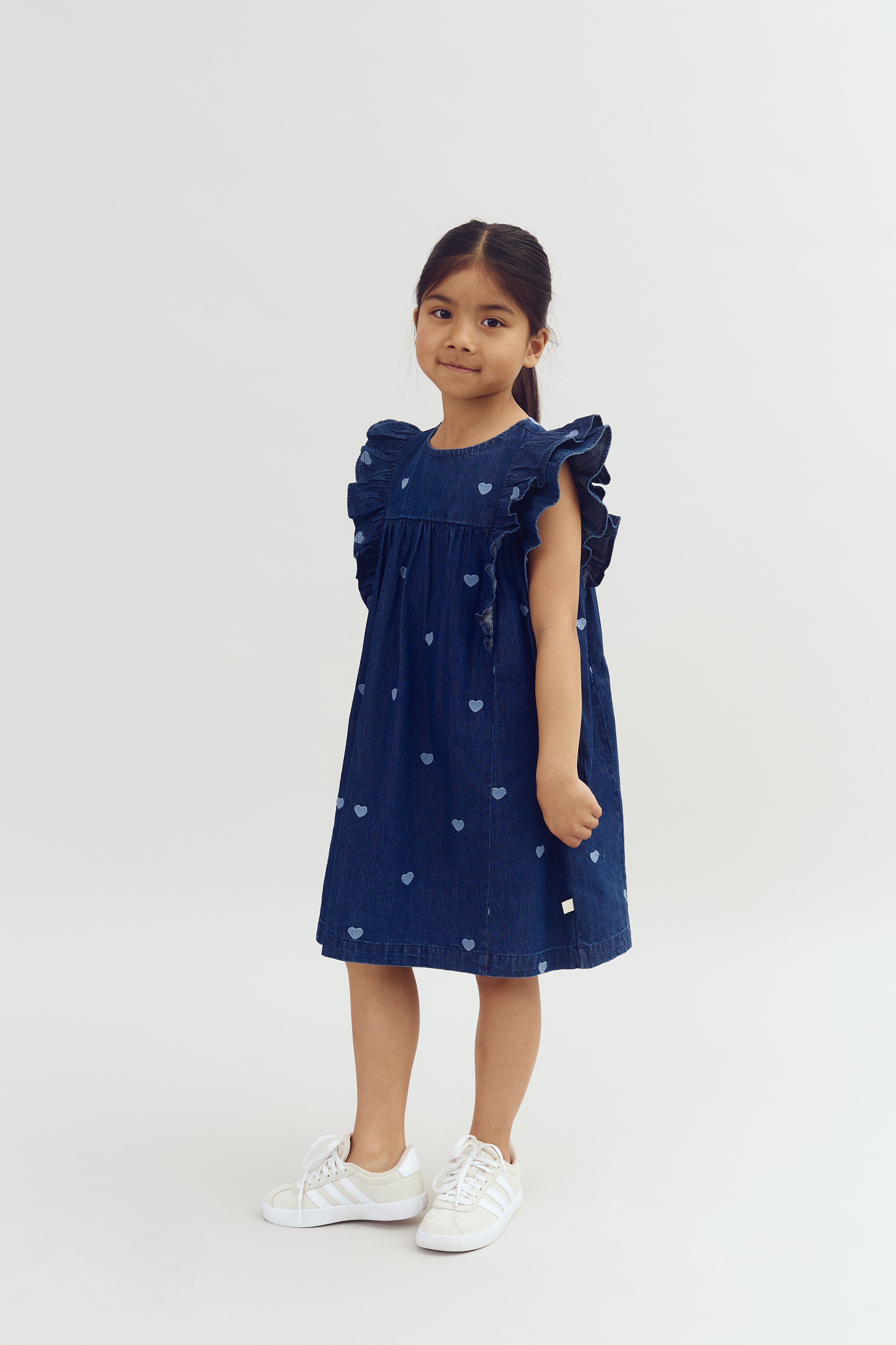 A young child stands confidently in a studio setting, wearing a blue dress with heart patterns and white sneakers, against a plain, light-colored background.
