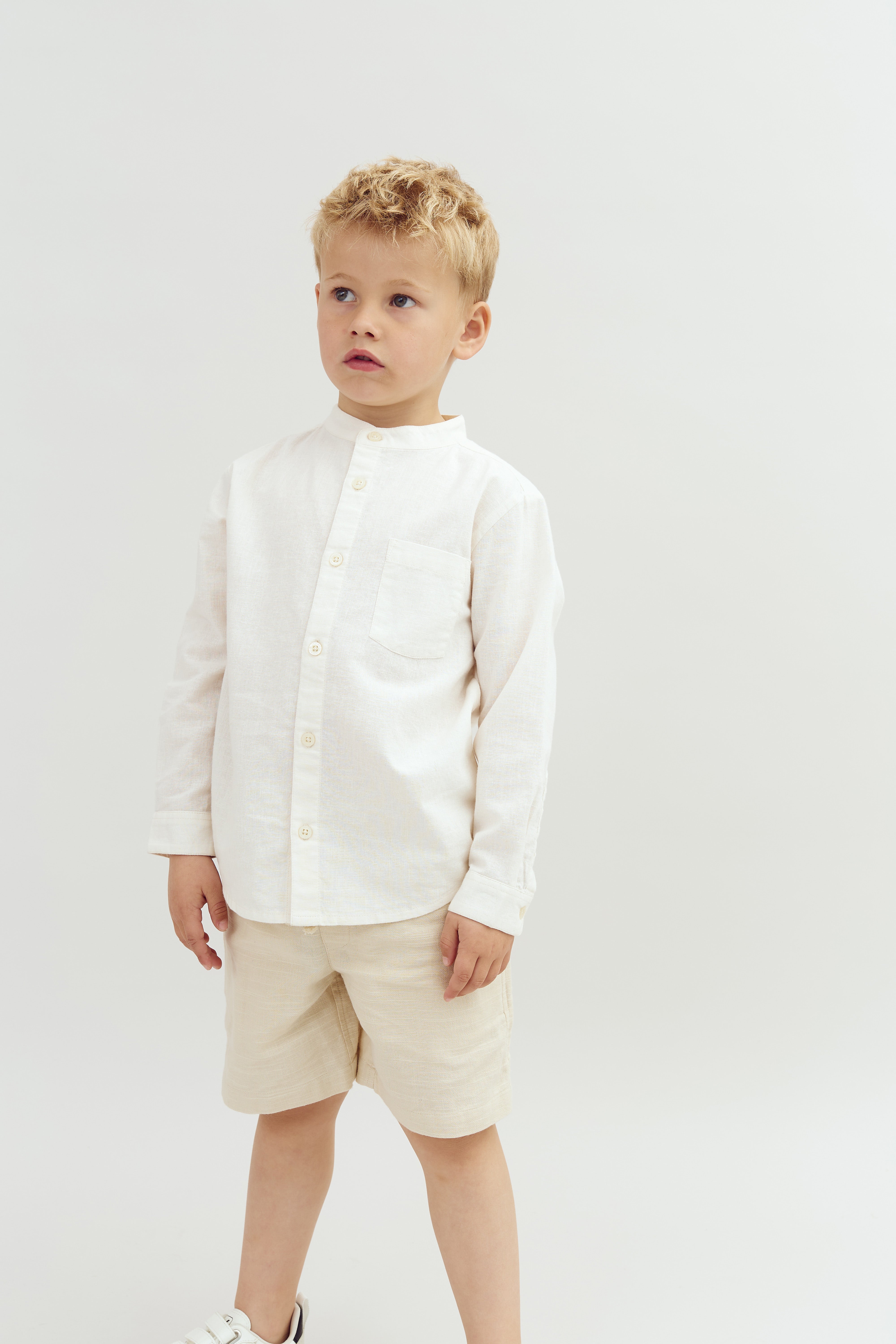 A young child stands wearing a white button-up shirt and beige shorts, looking slightly upwards in a neutral-colored studio setting.