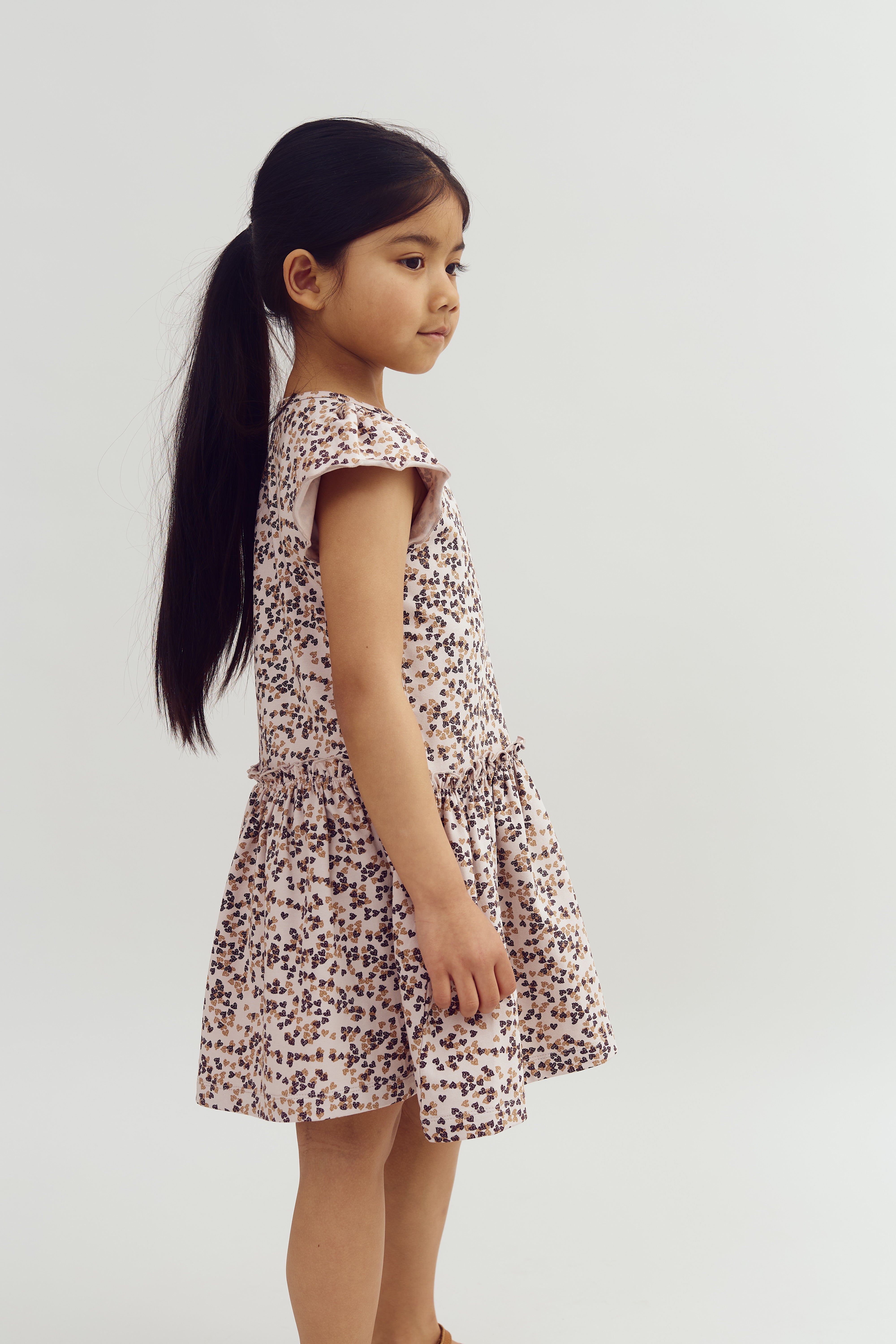 A young child stands sideways, wearing a floral-patterned dress. Her long, dark hair is tied back in a ponytail. The background is plain white.