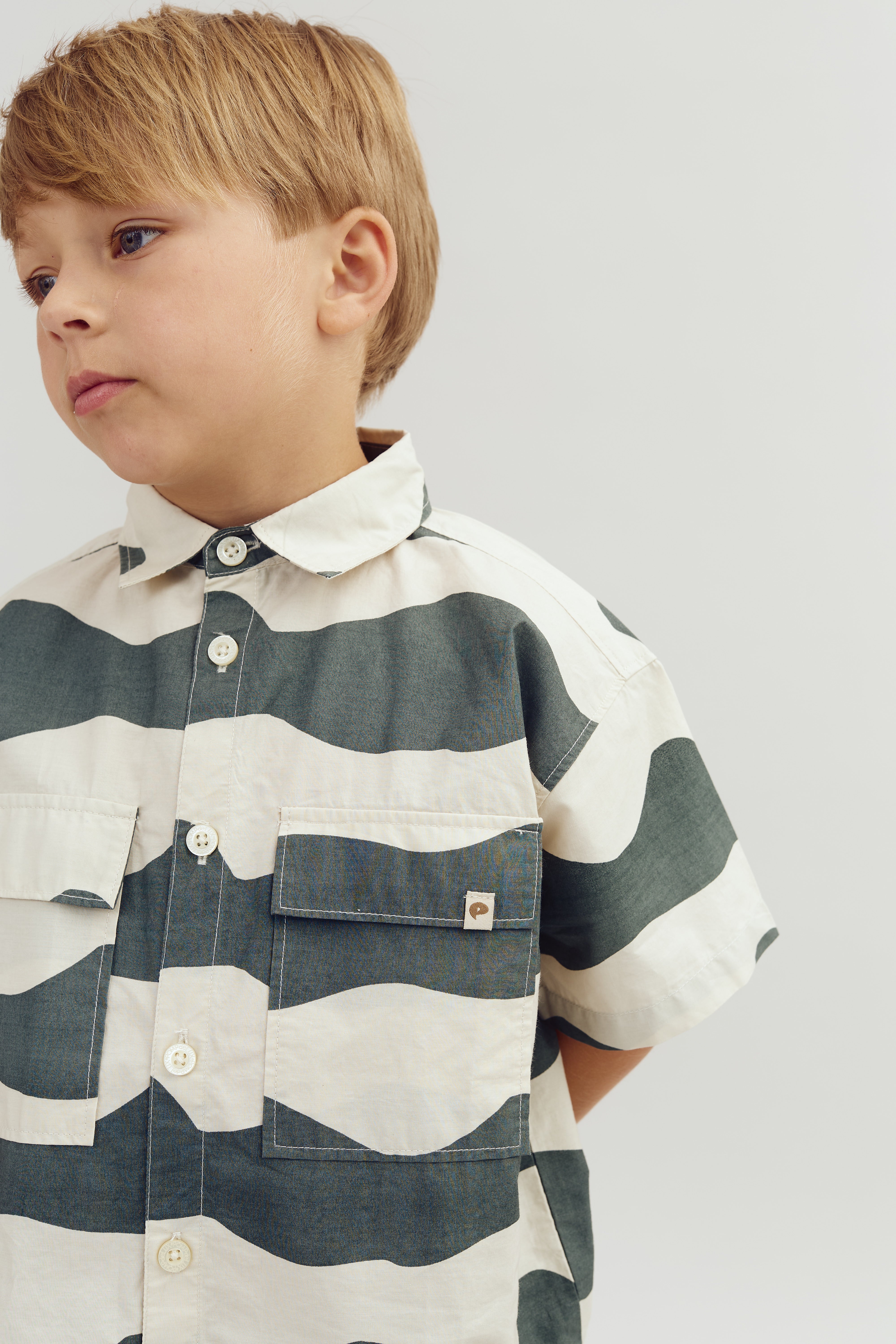 A young boy stands with a neutral expression, wearing a striped shirt with button-up detail, in front of a plain white background. A small logo is visible on the pocket.