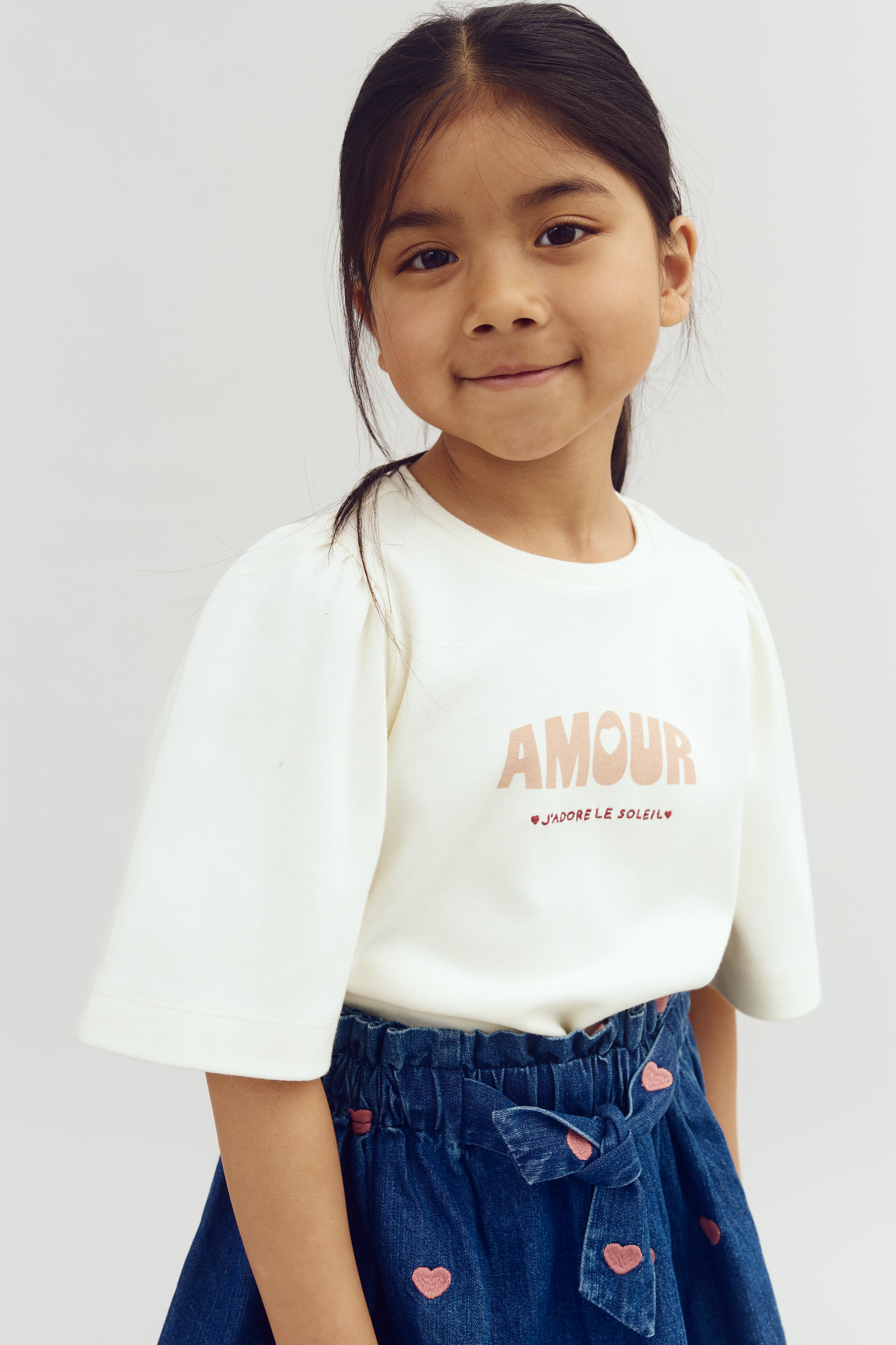 A smiling child wears a white shirt with “AMOUR” and “J’ADORE LE SOLEIL” printed on it, paired with a blue skirt adorned with small pink hearts, against a plain backdrop.
