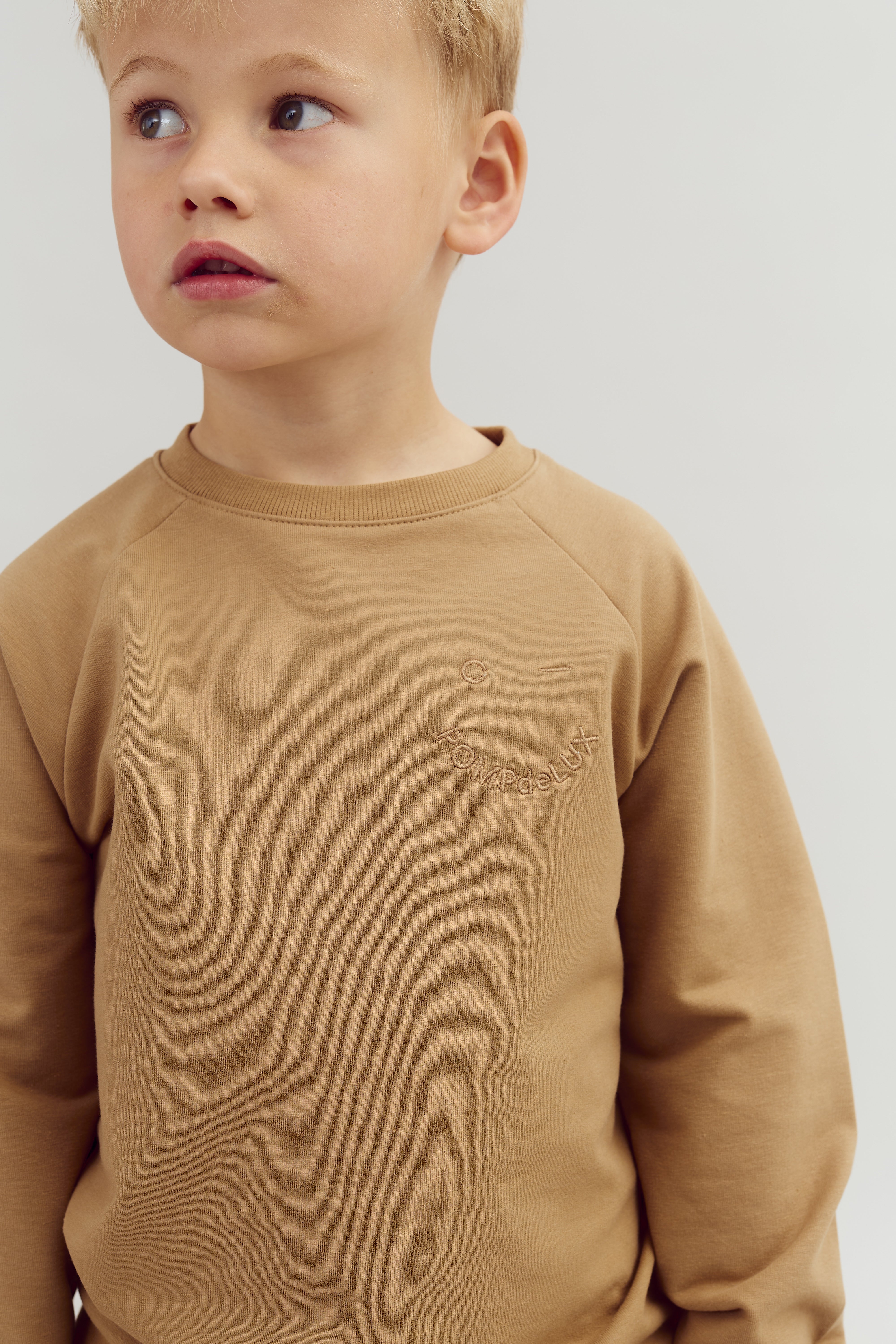 A young child is looking to the side, wearing a tan sweatshirt with the text "POMPdeLUX" embroidered on the front, standing against a plain background.