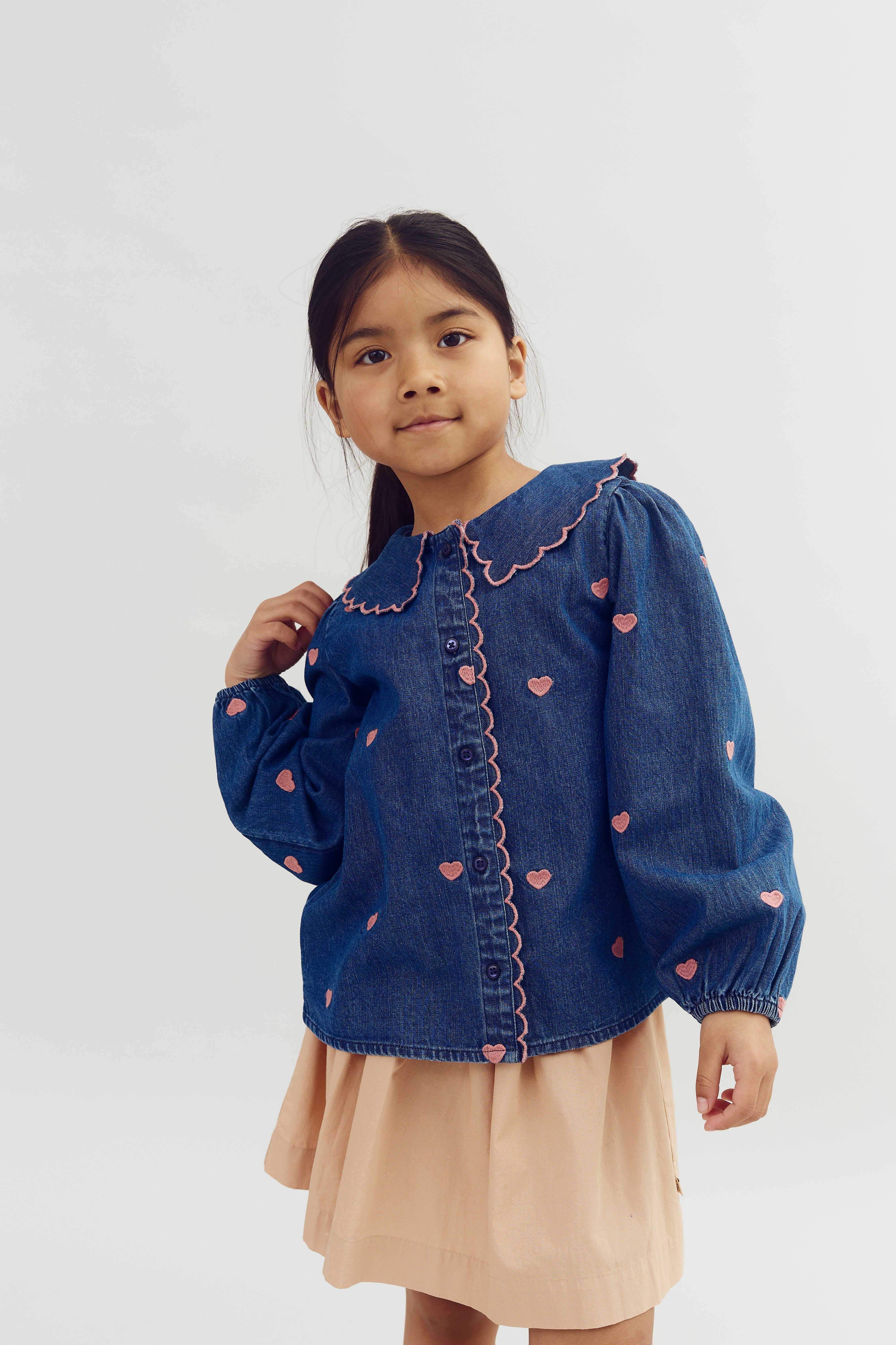 A child wearing a blue denim jacket with pink heart patterns and a beige skirt stands against a plain background. They have long dark hair tied back and are looking at the camera.