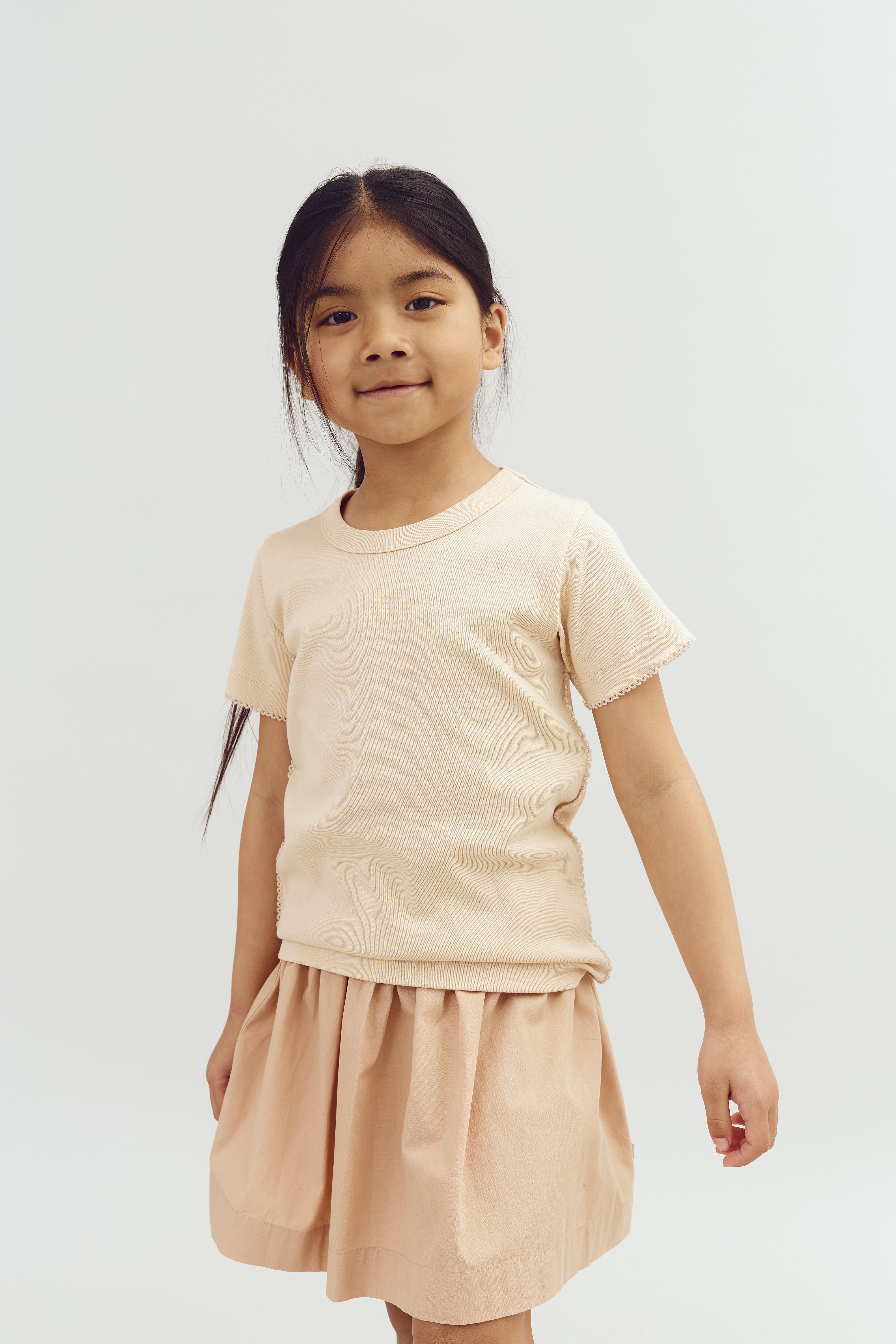 A young child stands and smiles wearing a cream-colored top and skirt, with long hair tied back, against a plain white background.
