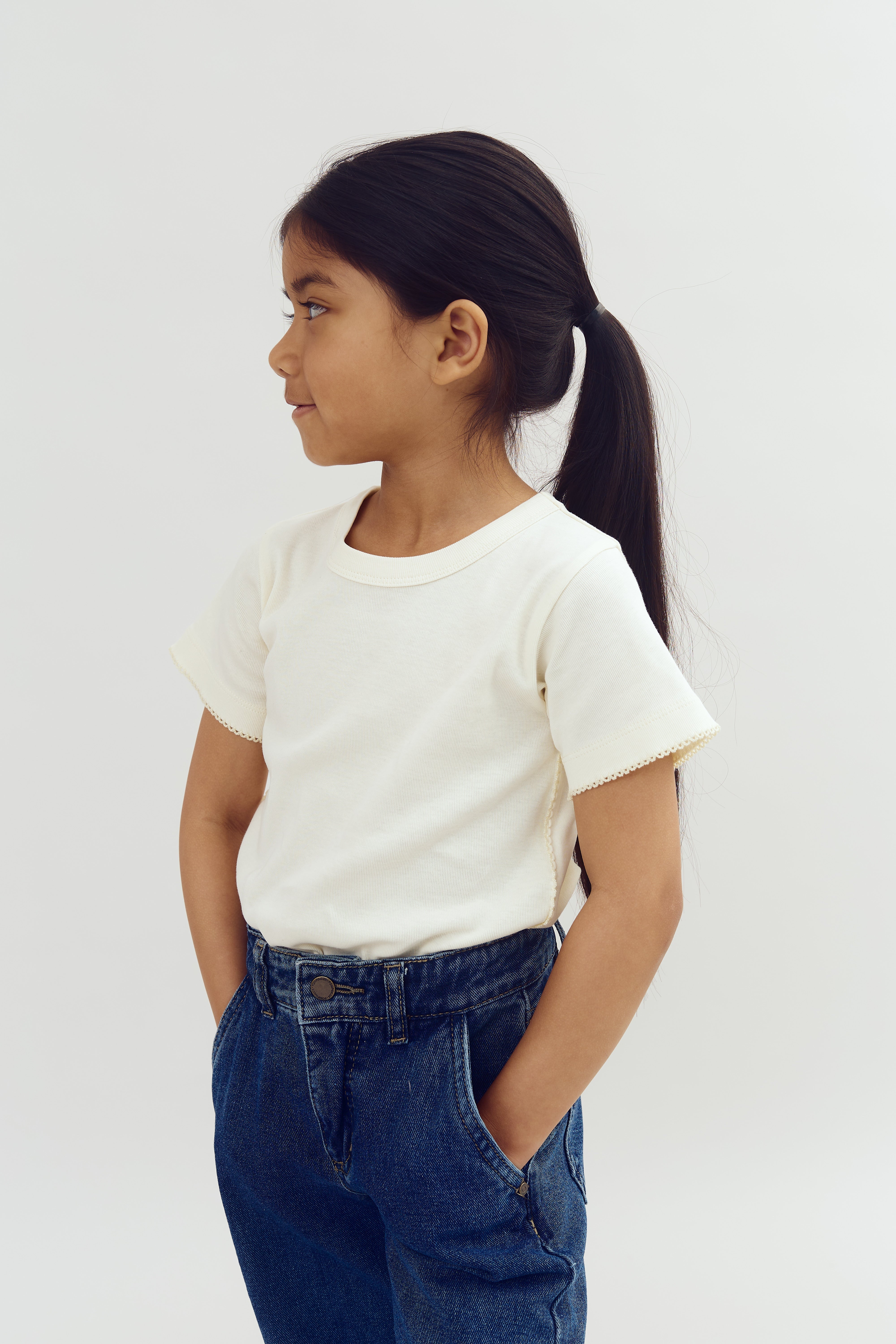 A child is standing, facing left, with hands in jean pockets. They wear a white shirt and have long hair tied back, against a plain light-colored background.