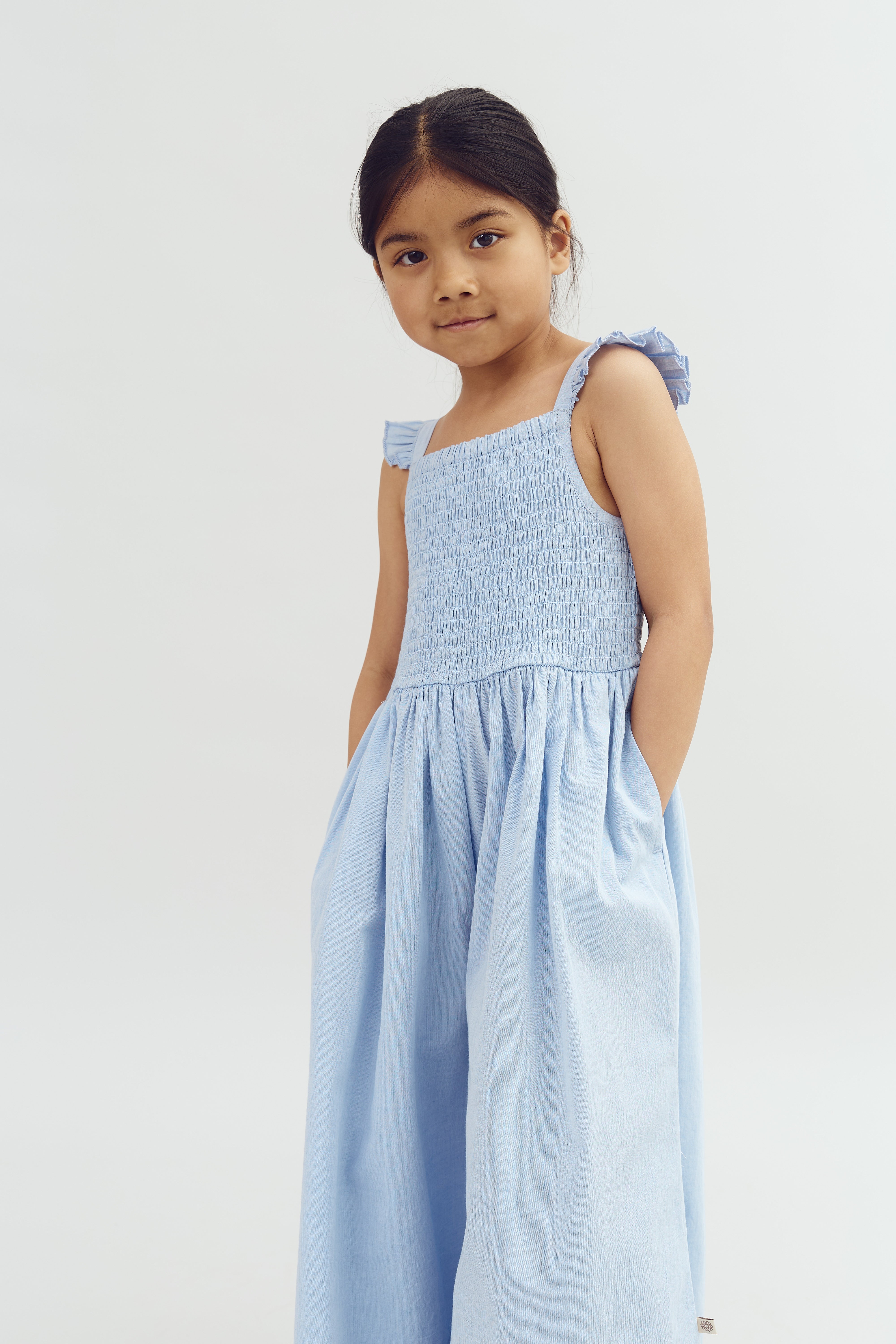 A young girl stands with hands in pockets, wearing a light blue, smocked dress with ruffled shoulder straps in a plain, white studio setting.