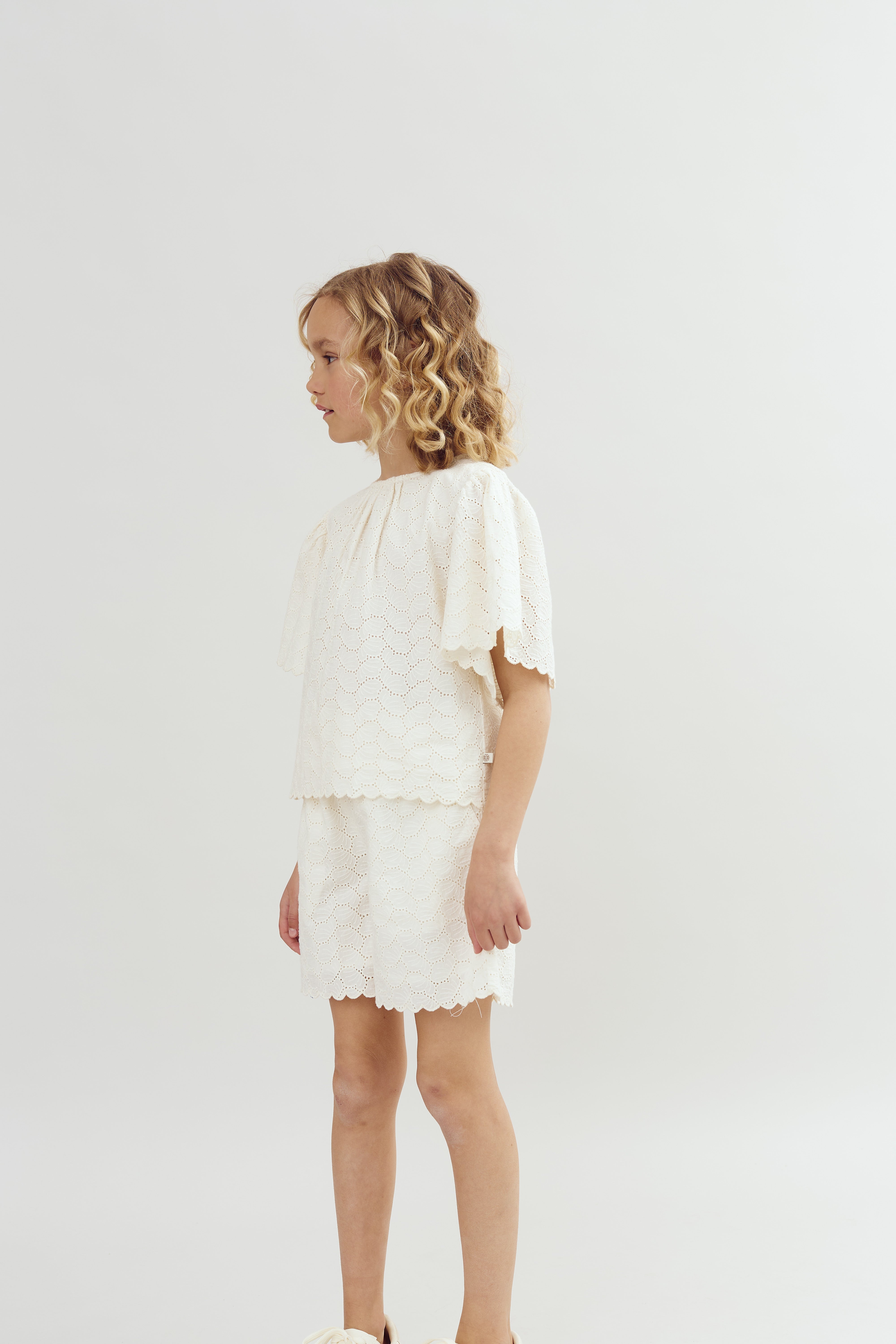 A child stands sideways wearing a white, scalloped, lace outfit with shorts and a top. The setting is a simple, neutral background, emphasizing the outfit's texture.