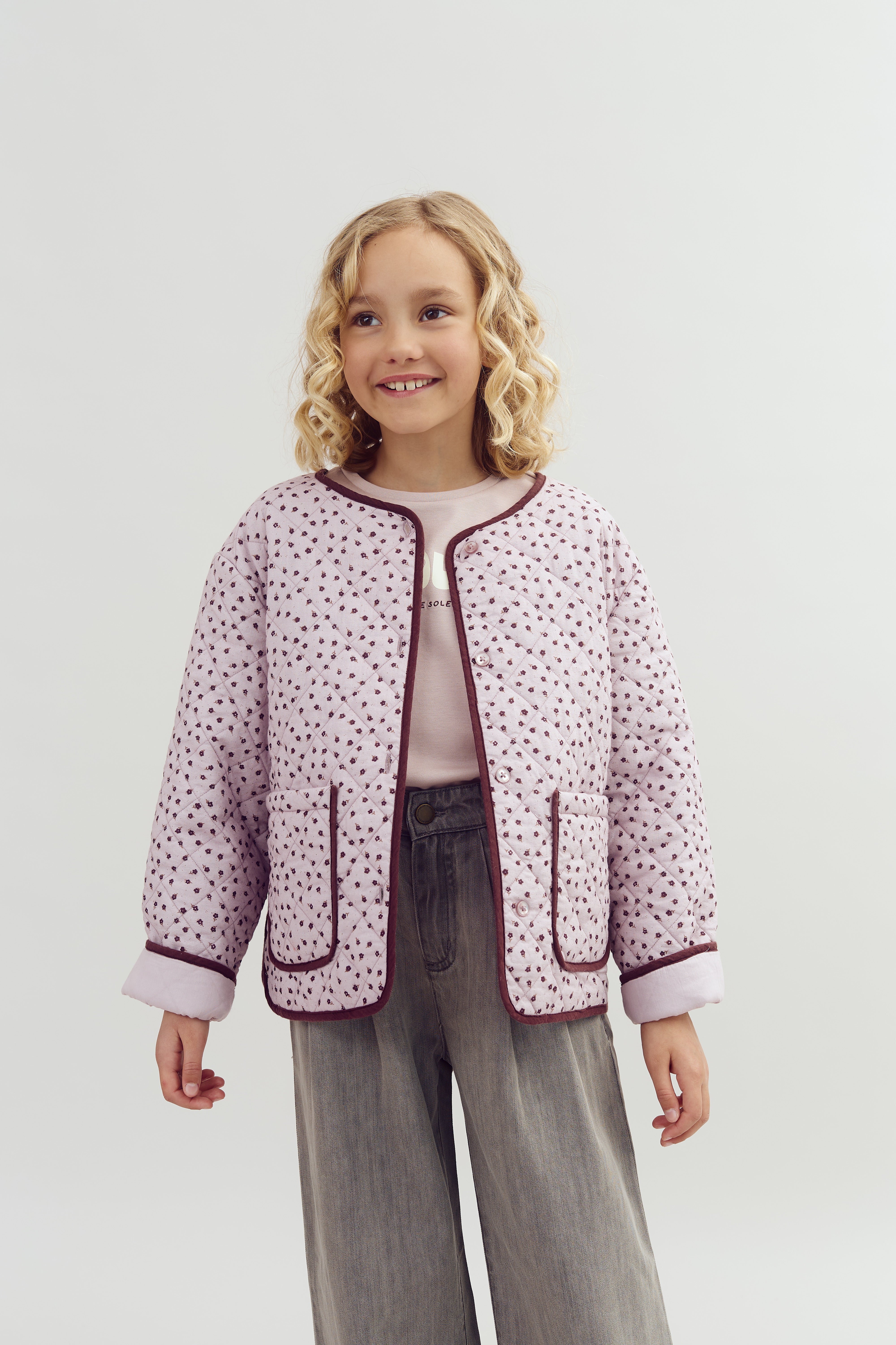 A child smiles while wearing a patterned quilted jacket over a pink shirt, with loose gray pants, set against a plain white backdrop.