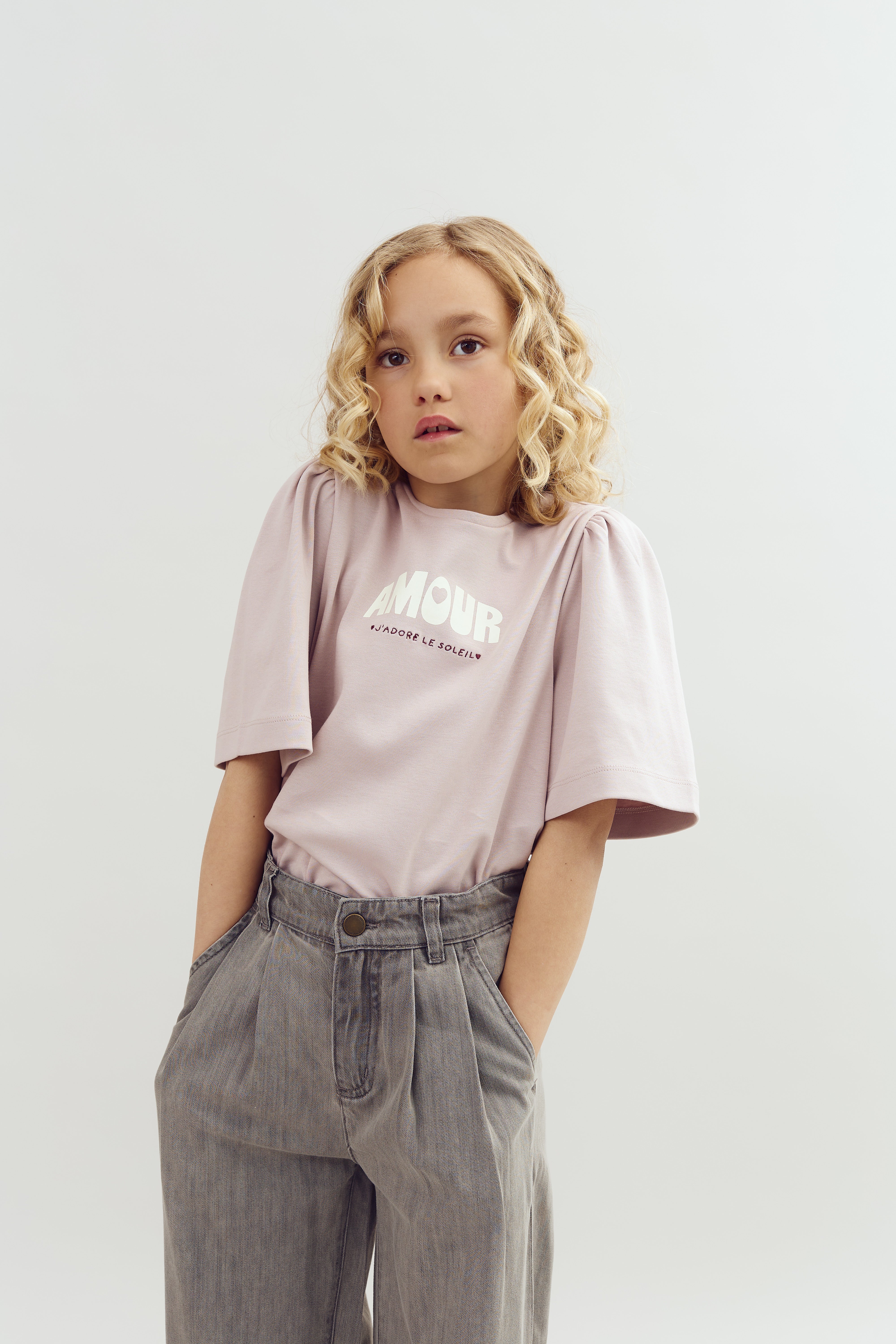 A child with curly blonde hair stands against a neutral background, wearing a light pink shirt with "AMOUR J'ADORE LE SOLEIL" printed on it, and grey trousers with hands in pockets.