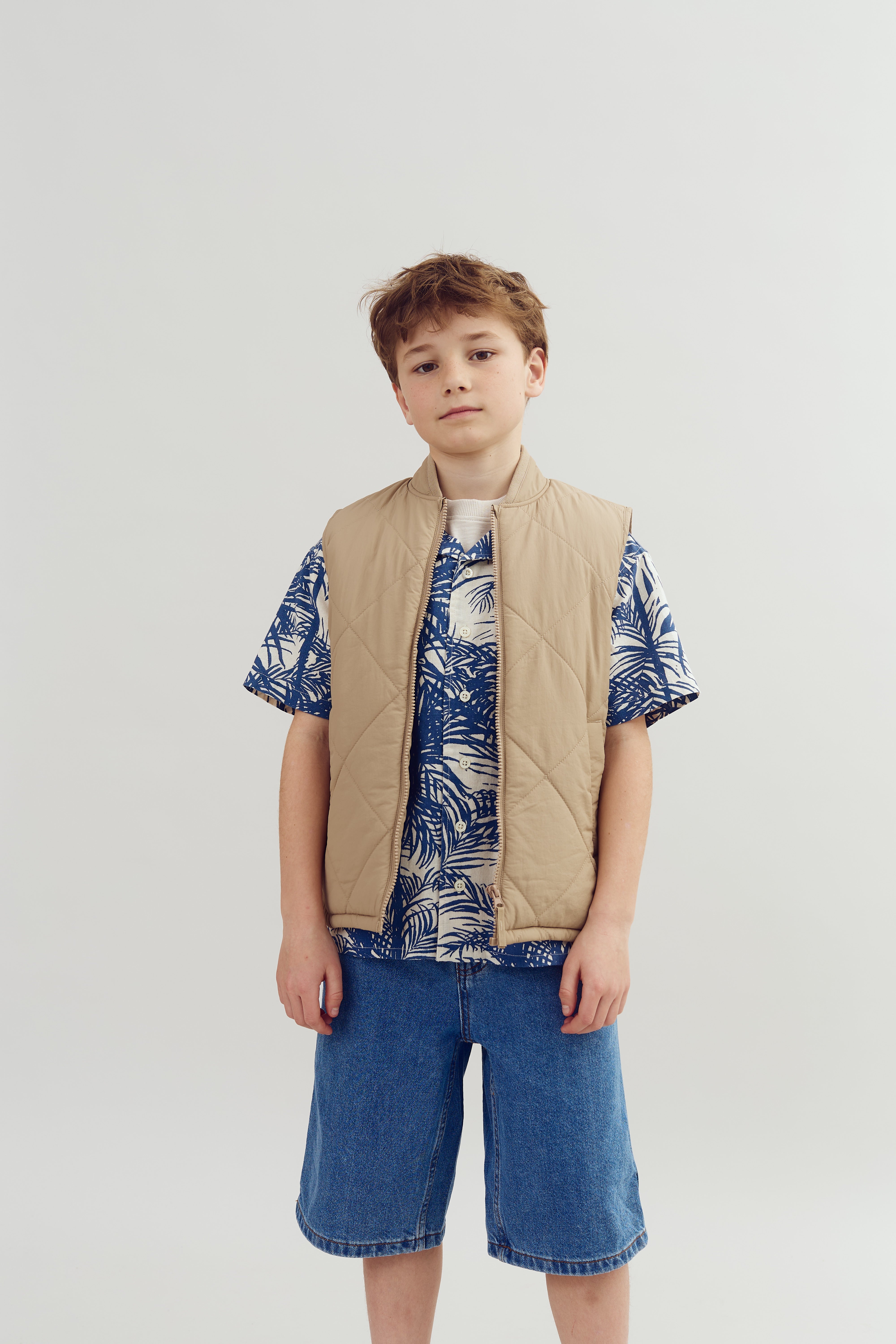 A young boy stands wearing a beige quilted vest over a blue patterned shirt and denim shorts against a plain white background, looking directly ahead.