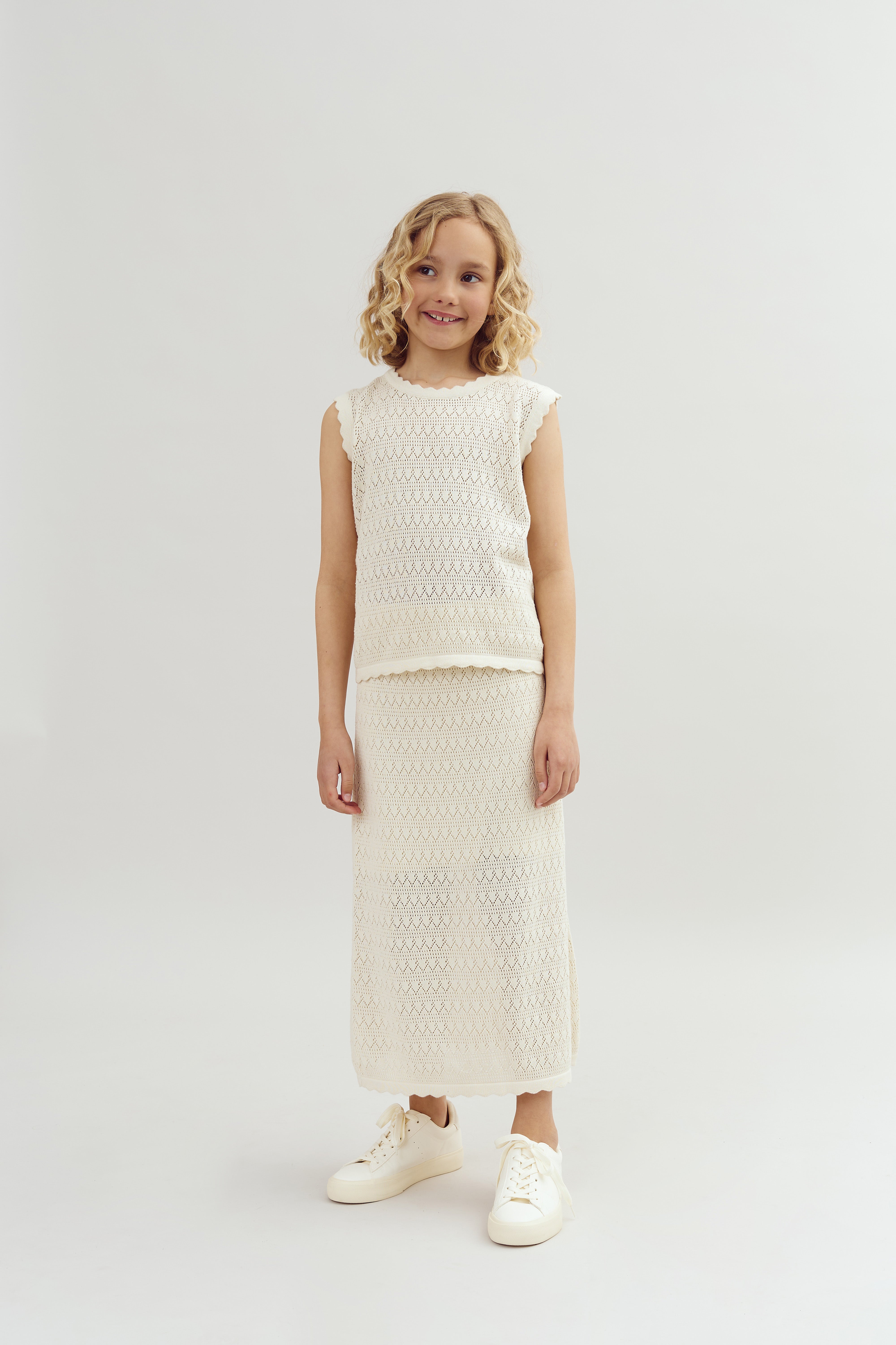 A young girl stands smiling, wearing a cream-colored crocheted top and skirt with white sneakers, against a plain, light gray background. Her hair is styled in loose curls.