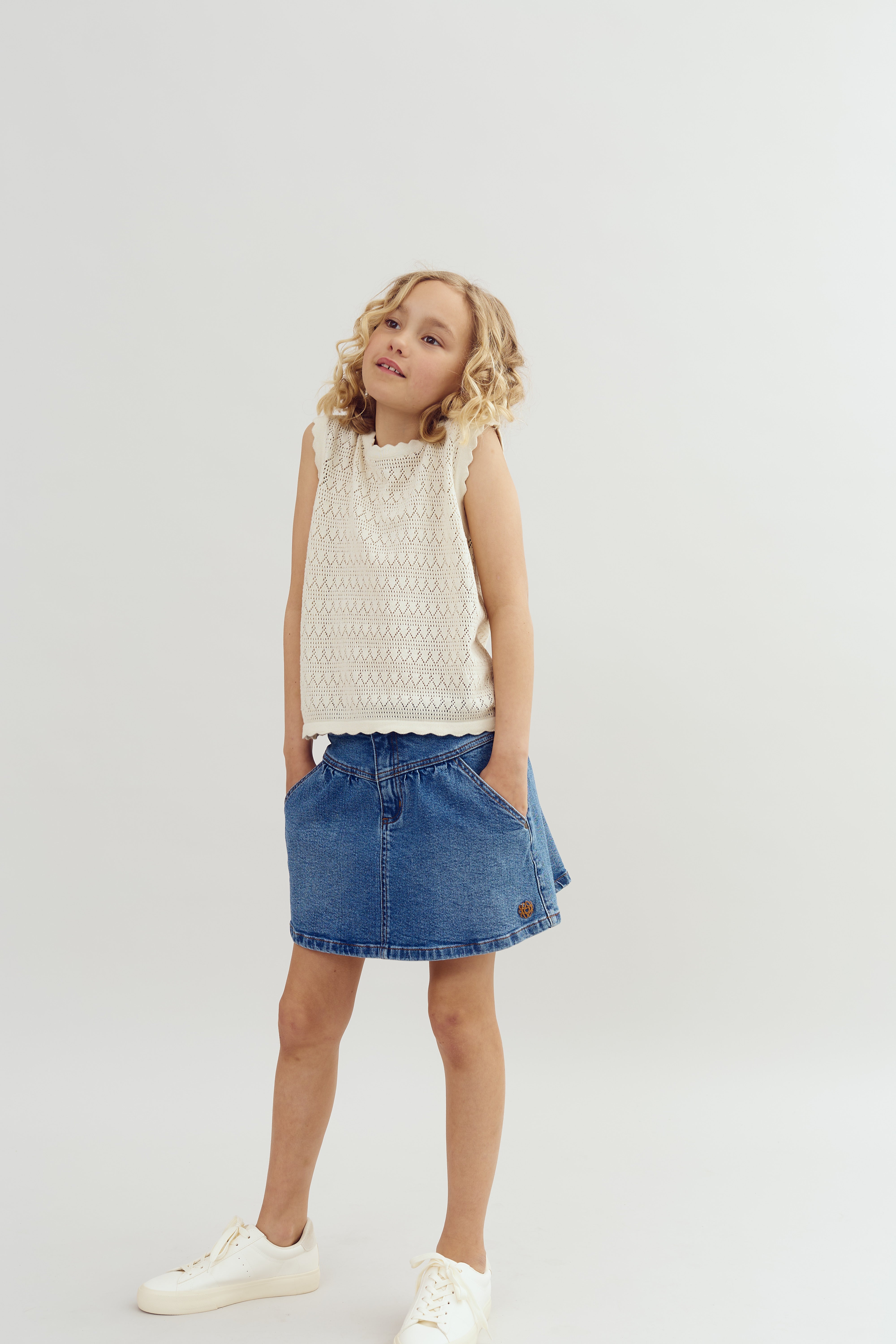 A young girl with curly hair stands, hands in pockets, wearing a white sleeveless top and blue denim skirt, against a plain white background.