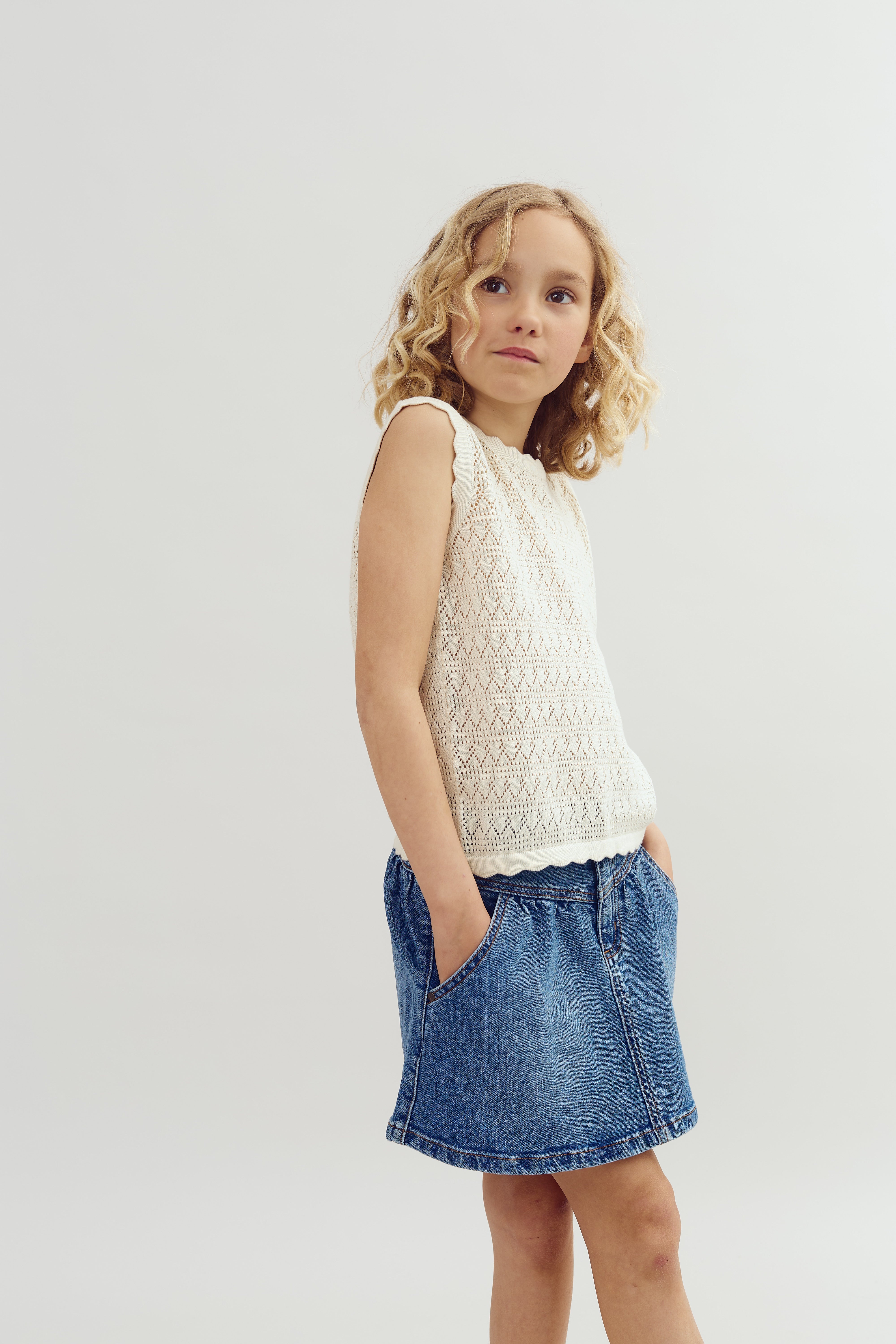 A young girl stands with hands in pockets, wearing a sleeveless white knit top and denim skirt, against a plain light background.