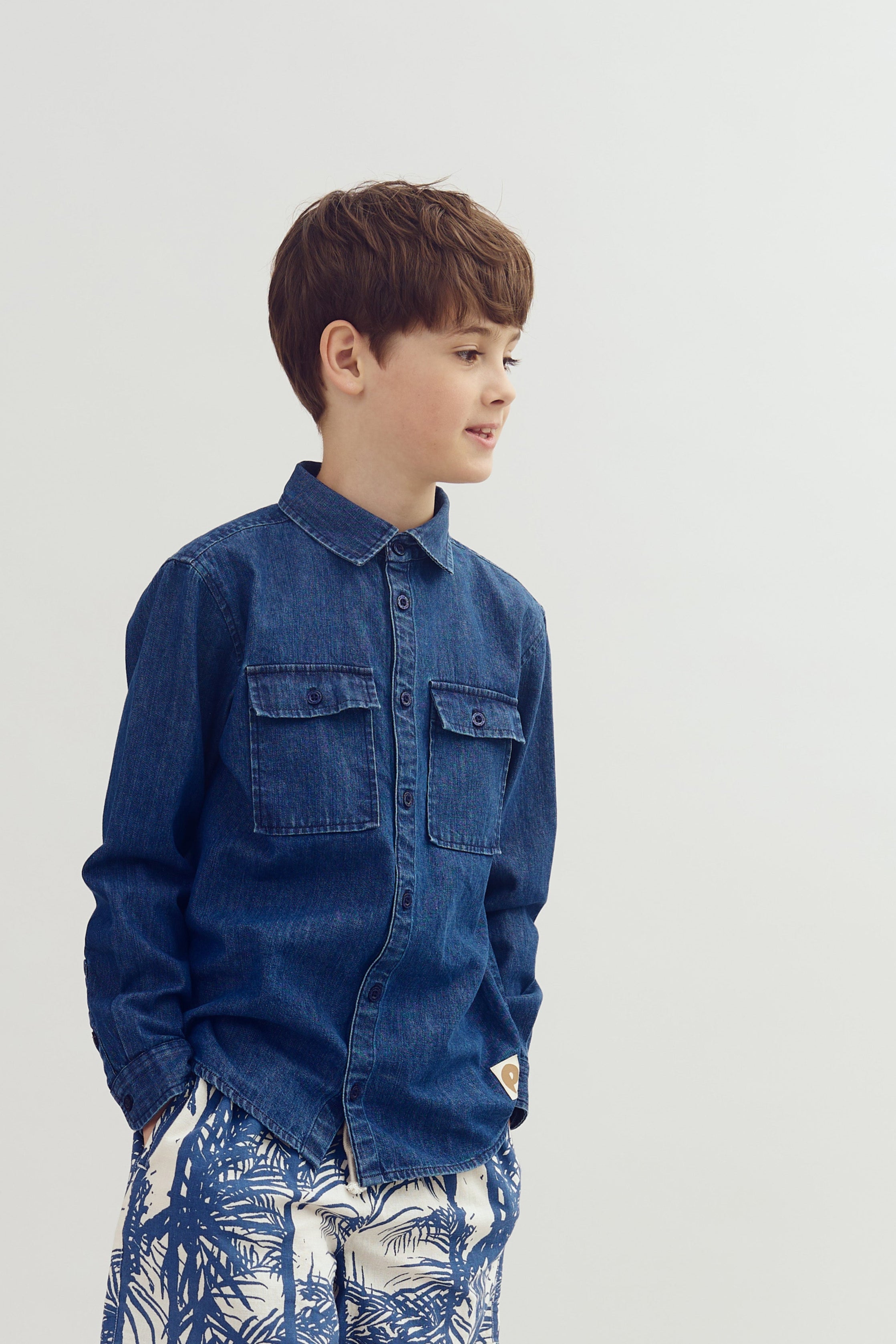 A young boy stands, hands in pockets, wearing a denim shirt and patterned shorts. He faces right against a plain white background, appearing thoughtful and relaxed.