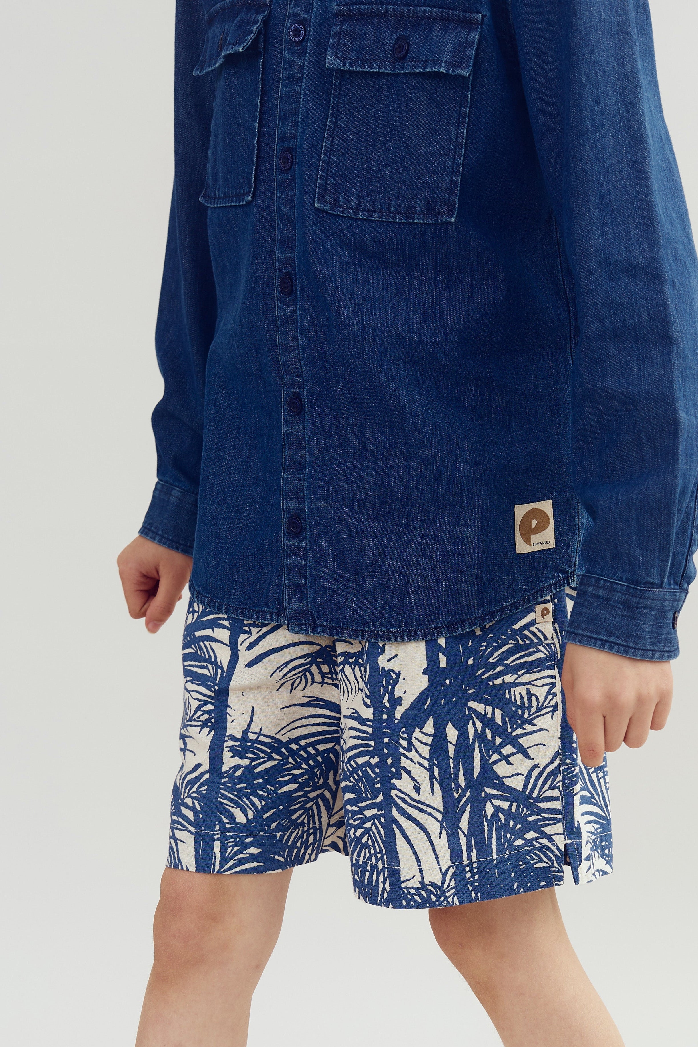 A child wears a blue denim shirt and patterned shorts with leaf designs, standing against a plain background. The shirt has pockets and a label with a capital "P."