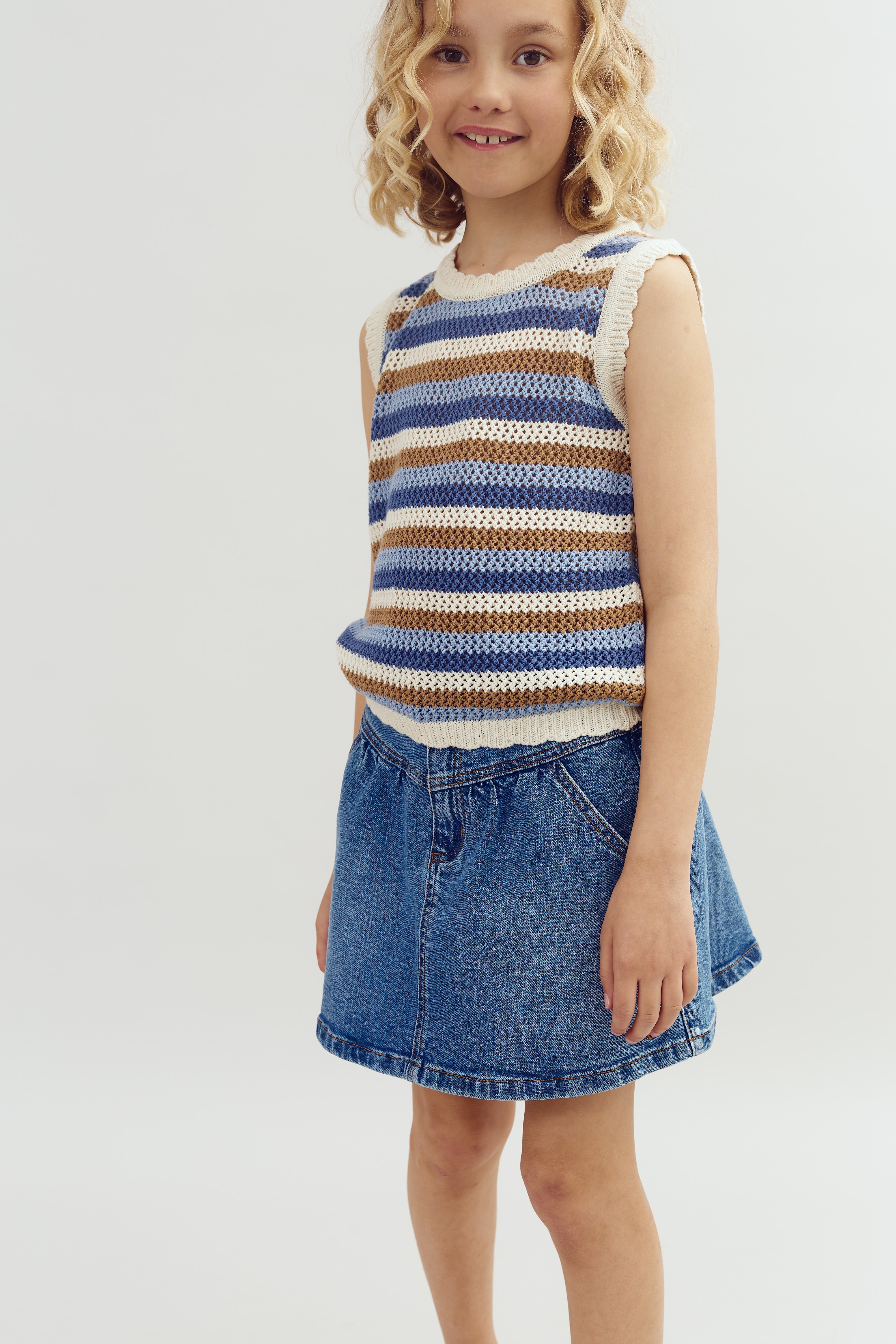A child stands smiling, wearing a sleeveless, blue and brown striped sweater and a denim skirt, against a plain white background.