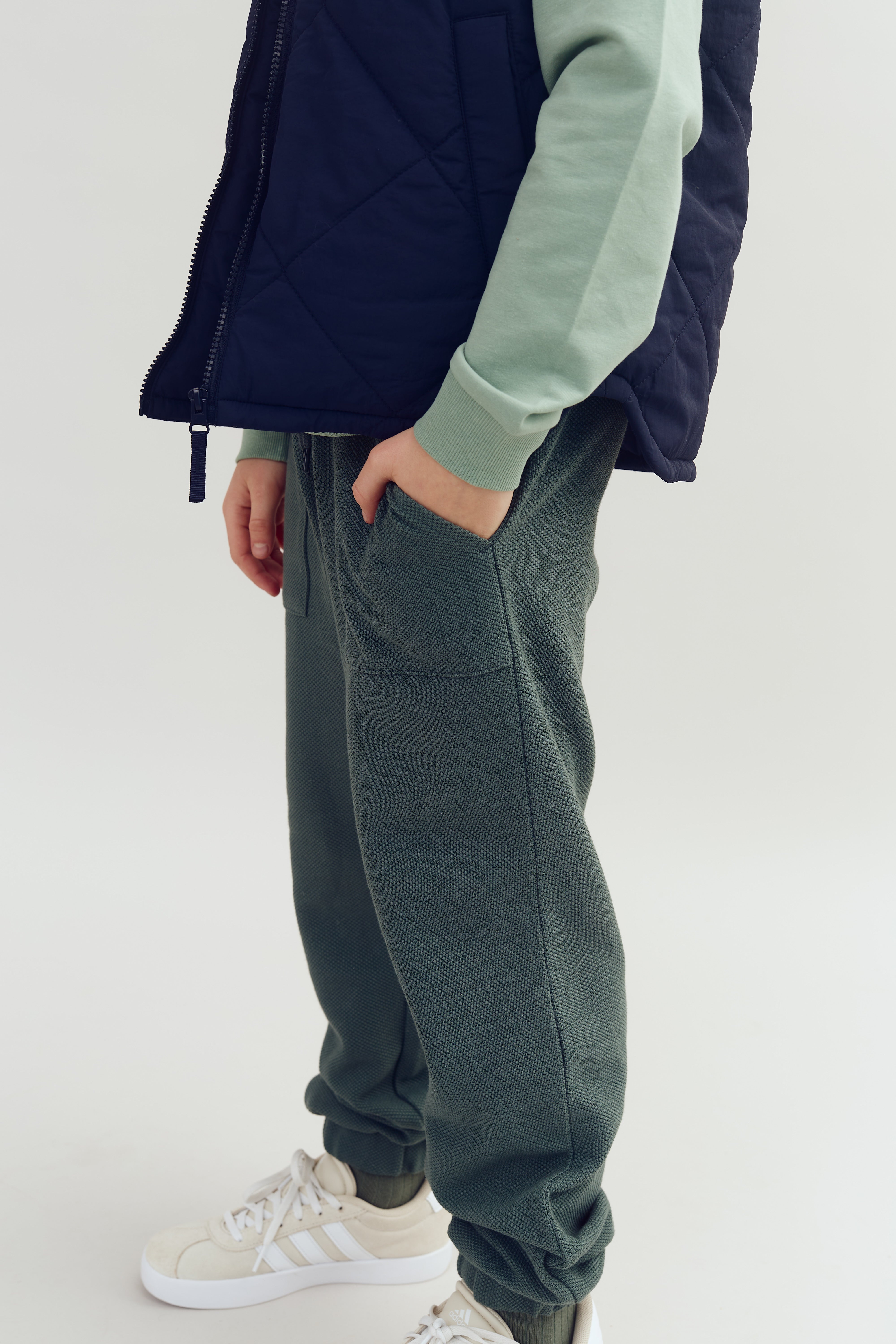 Person wearing a navy vest, mint green long-sleeved shirt, and dark green textured pants with hand in pocket. Standing on a light background, they wear white sneakers with visible stripes.