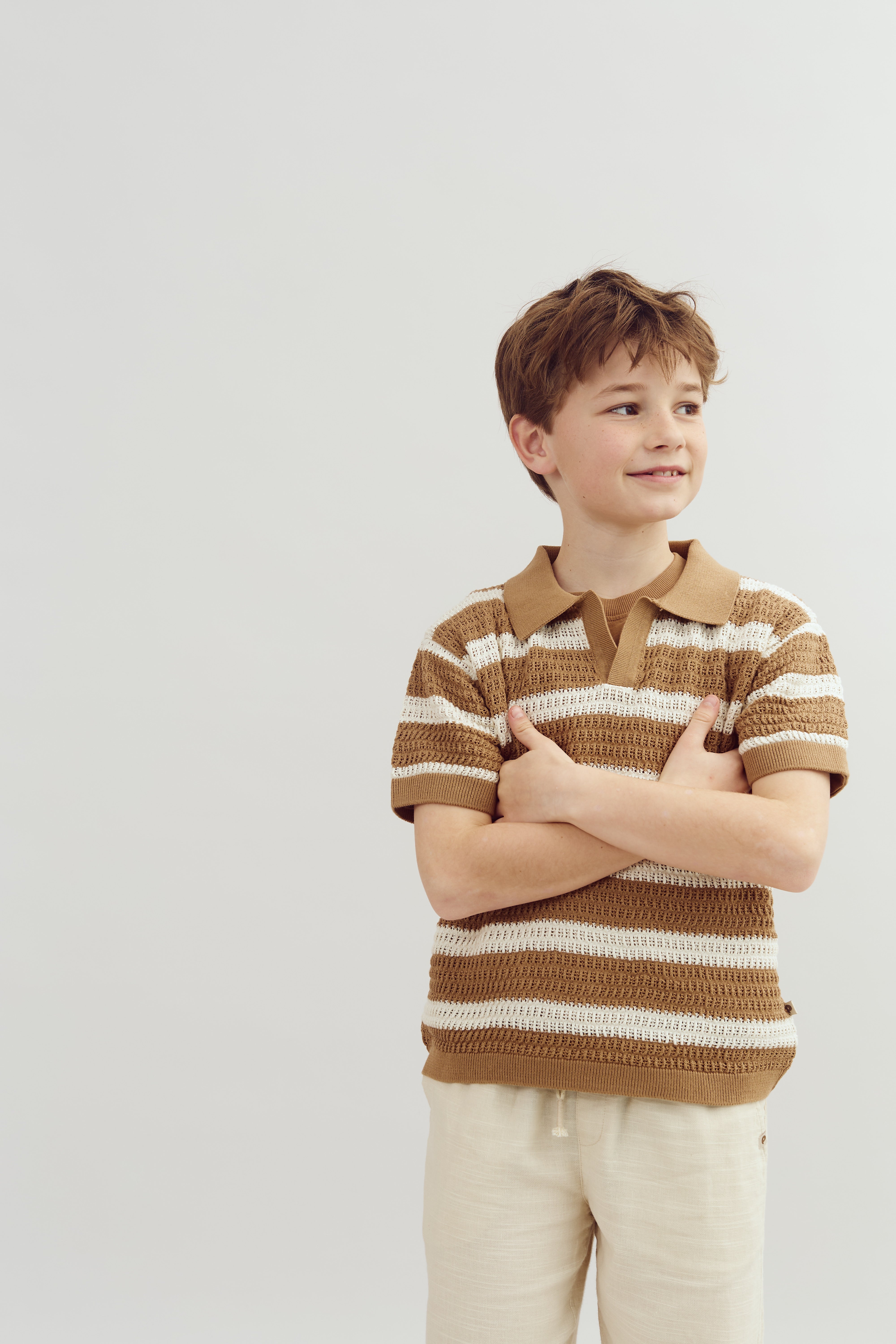 A child stands smiling with arms crossed, wearing a brown and white striped shirt. He is in a light, neutral-colored environment.