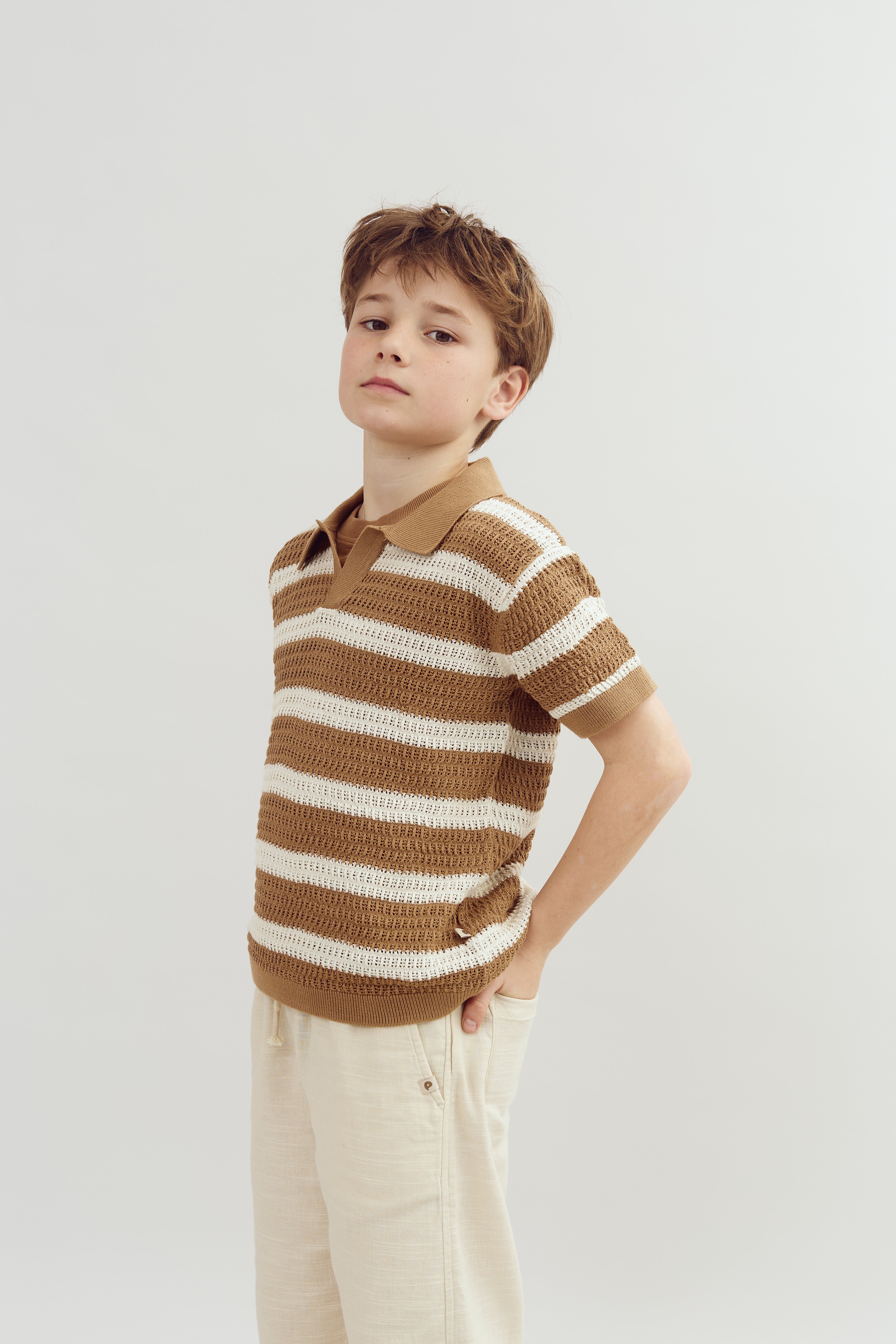 A young child stands confidently, wearing a brown and white striped shirt and beige pants, with hands in pockets against a plain, light background.