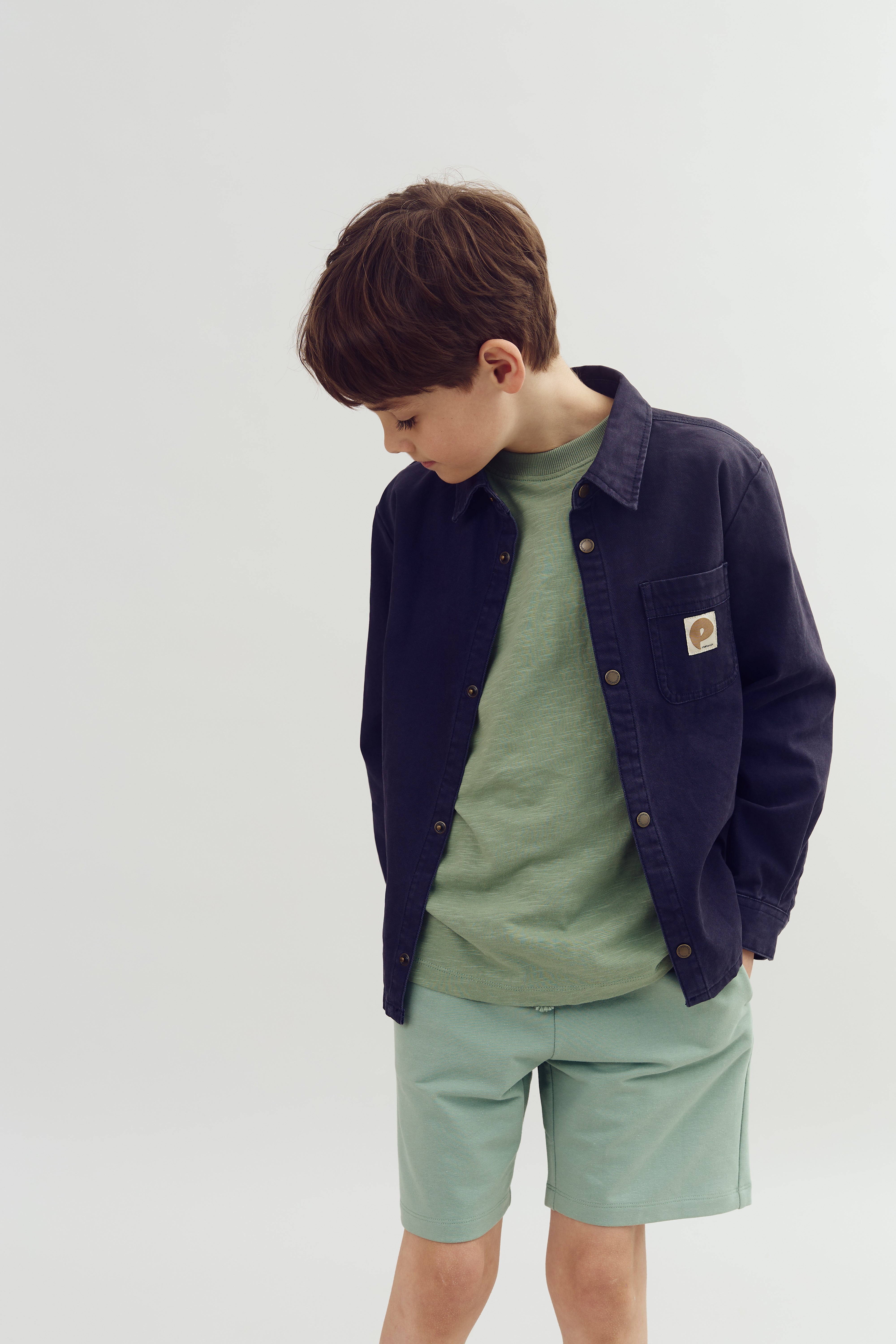 A child stands, looking down, wearing a blue jacket and green shirt with matching shorts in a plain, light-colored setting. The jacket has a small emblem on the pocket.