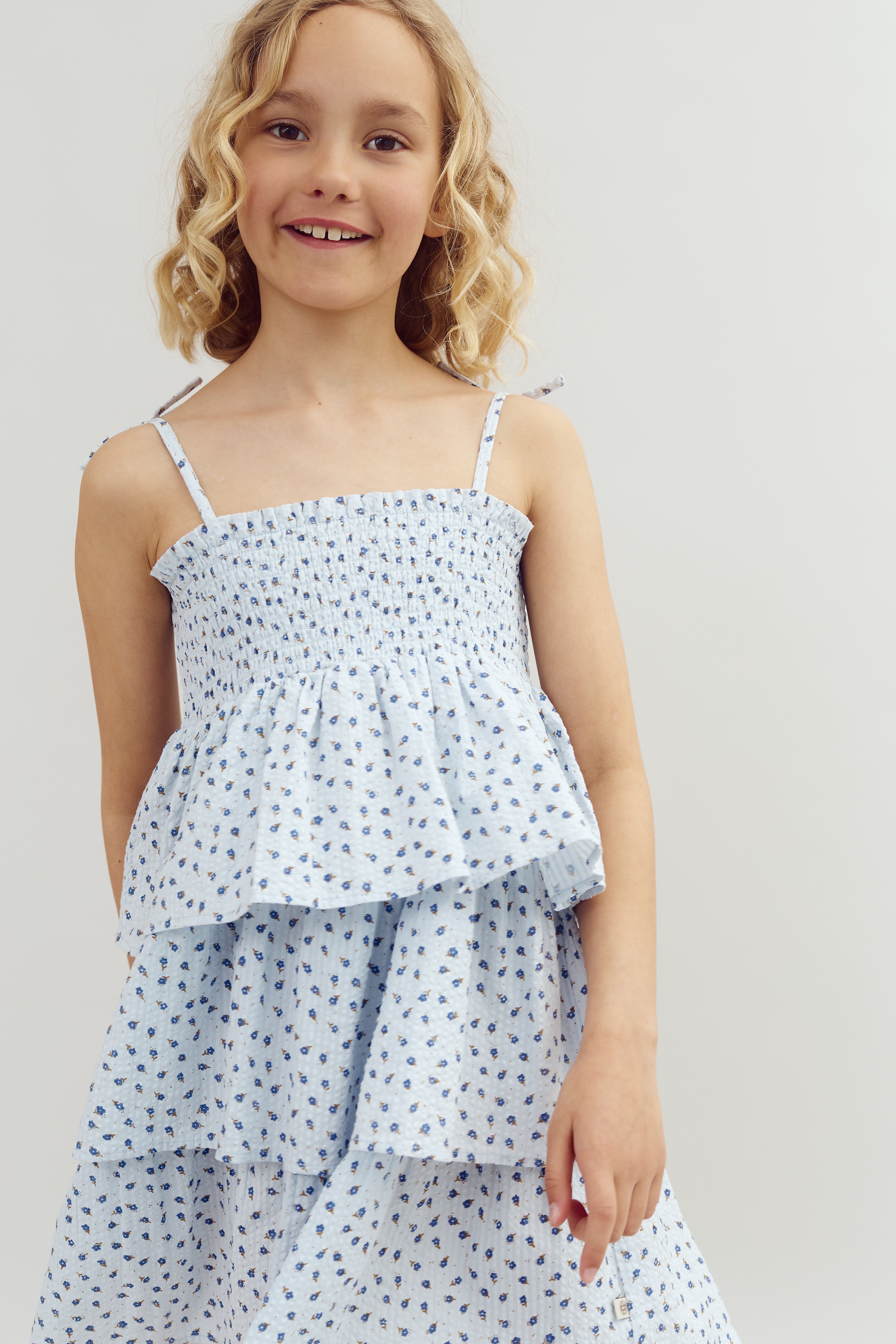 A smiling child in a light blue, floral dress stands against a plain, light-colored background. The dress features small blue floral patterns and ruffled tiers.